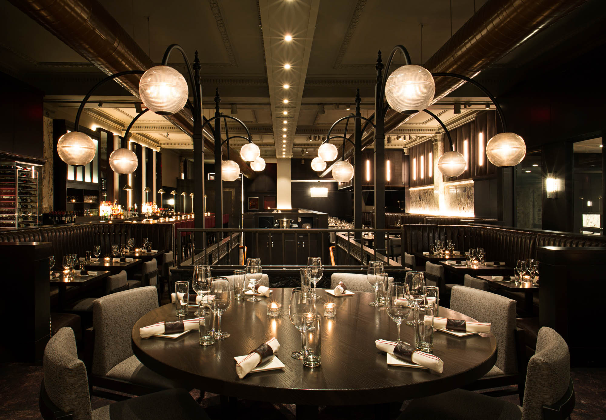 A wide shot of Boston Chops DTX, looking down the length of the restaurant and towards the front door, with elegant light fixtures and a long bar in view.