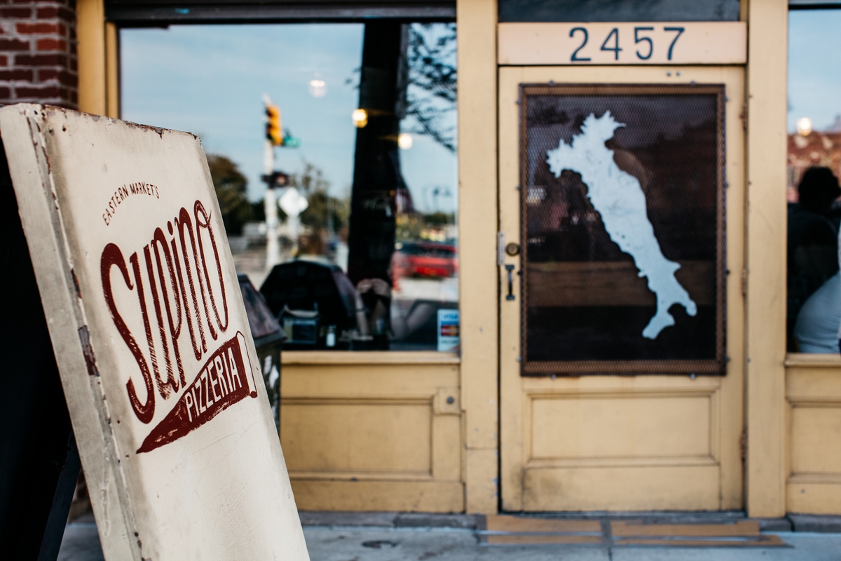 A sandwich board reads Supino Pizza in front of the restaurant. The window to the door features a decal in the shape of Italy’s “boot.”