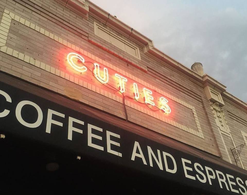 Cuties Coffee neon sign hangs on a brick building over a coffee shop.