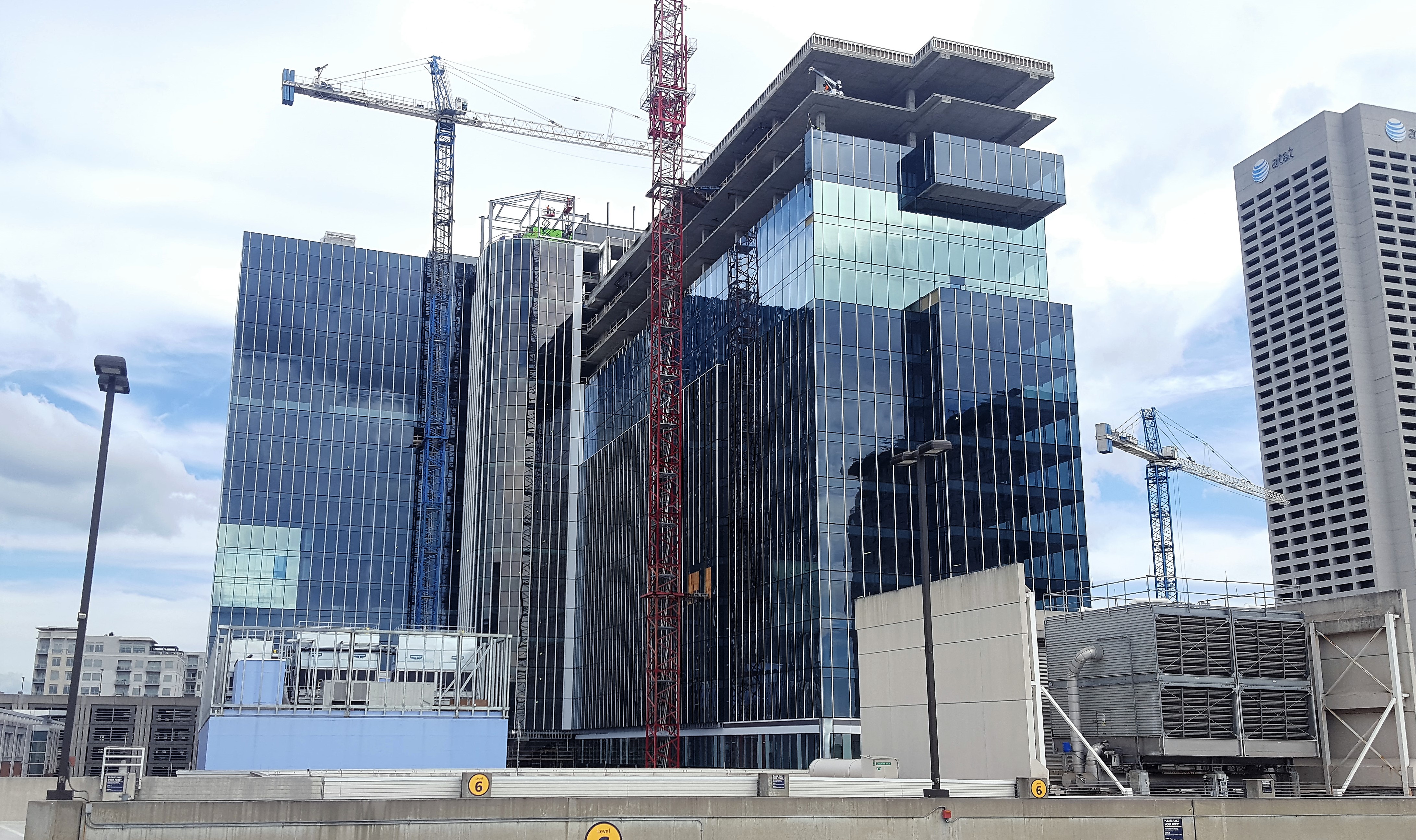 A glassy, blue, L-shaped office tower is under construction and surrounded by cranes.