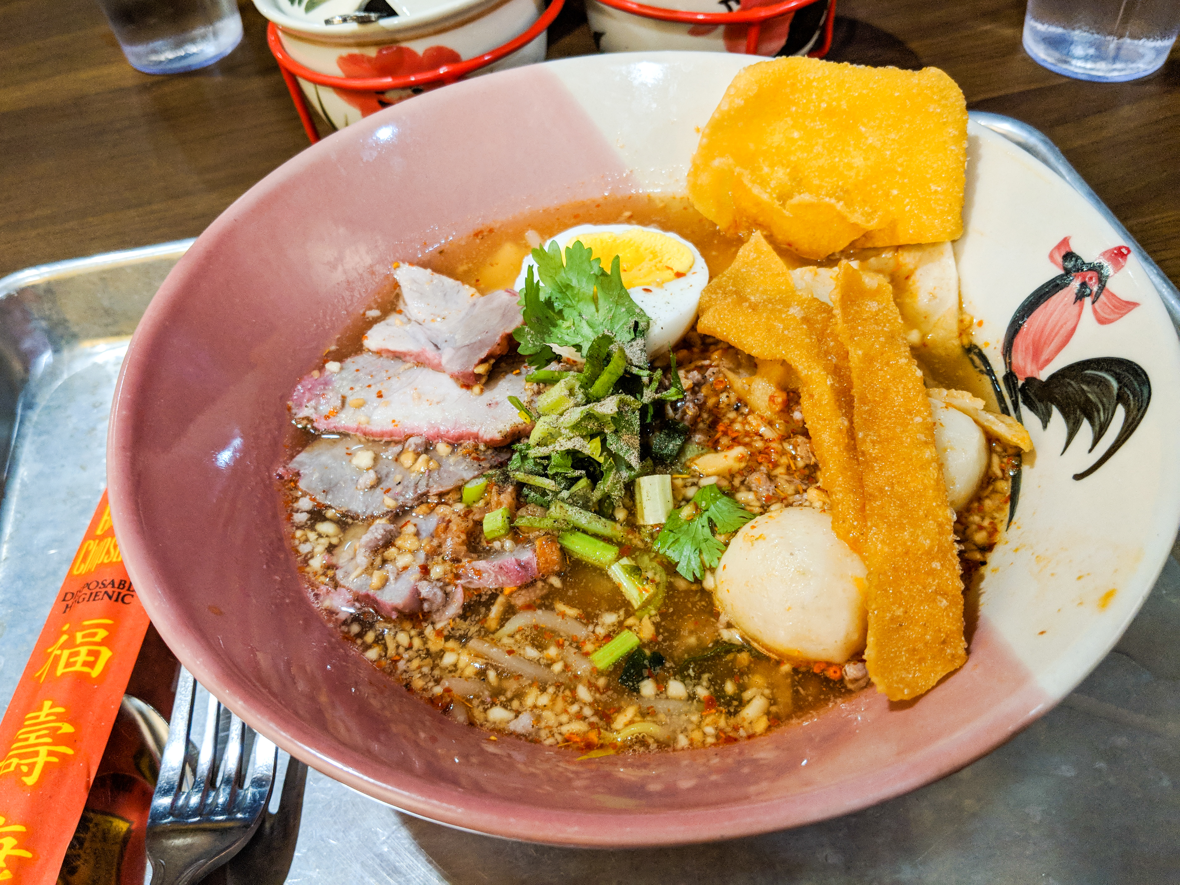 Soup in a Thai-style bowl decorated with a rooster. There are pink barbecue pork slices in the soup, as well as an egg, ground pork, ground peanuts, crispy wonton strips, and more. The bowl sits on a metal tray.