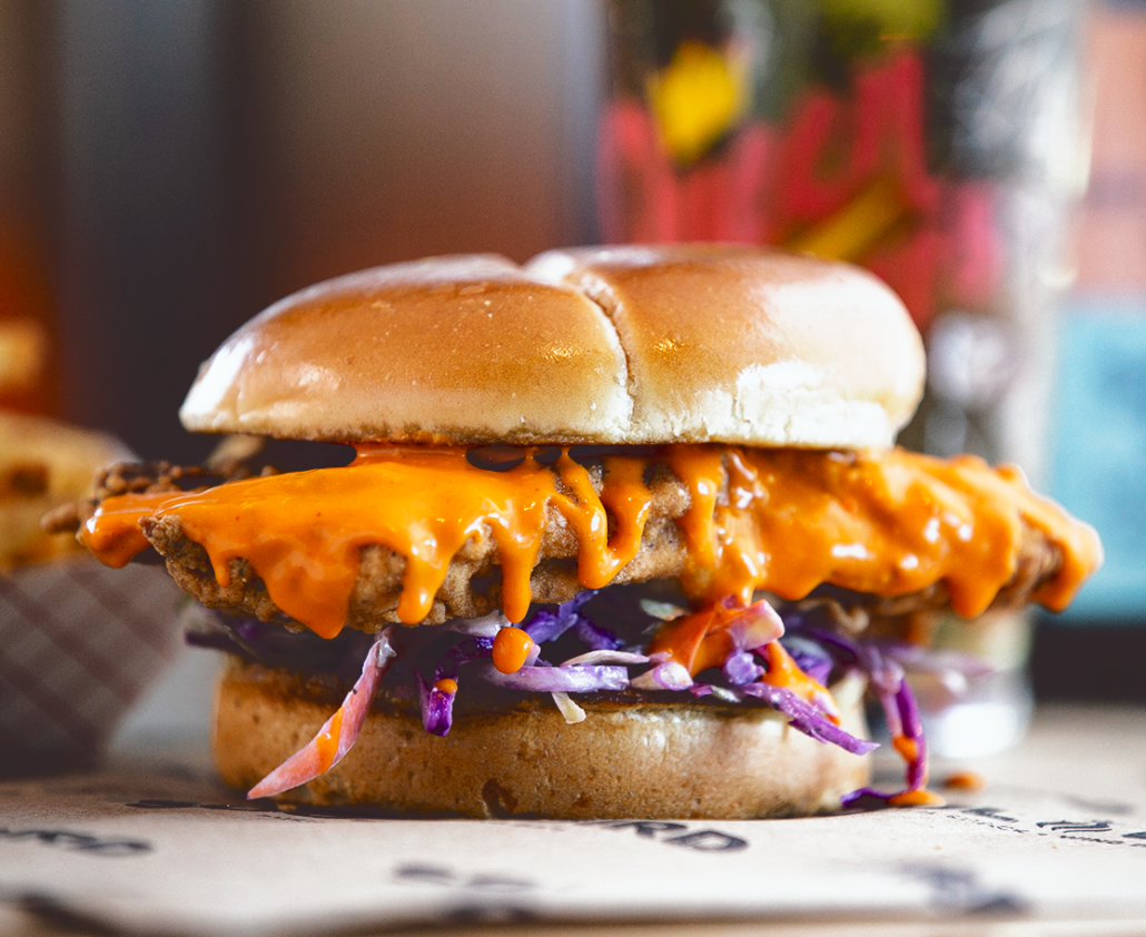A chicken sandwich is covered in an orange sauce that drips off the sides and sits on purple cabbage. 