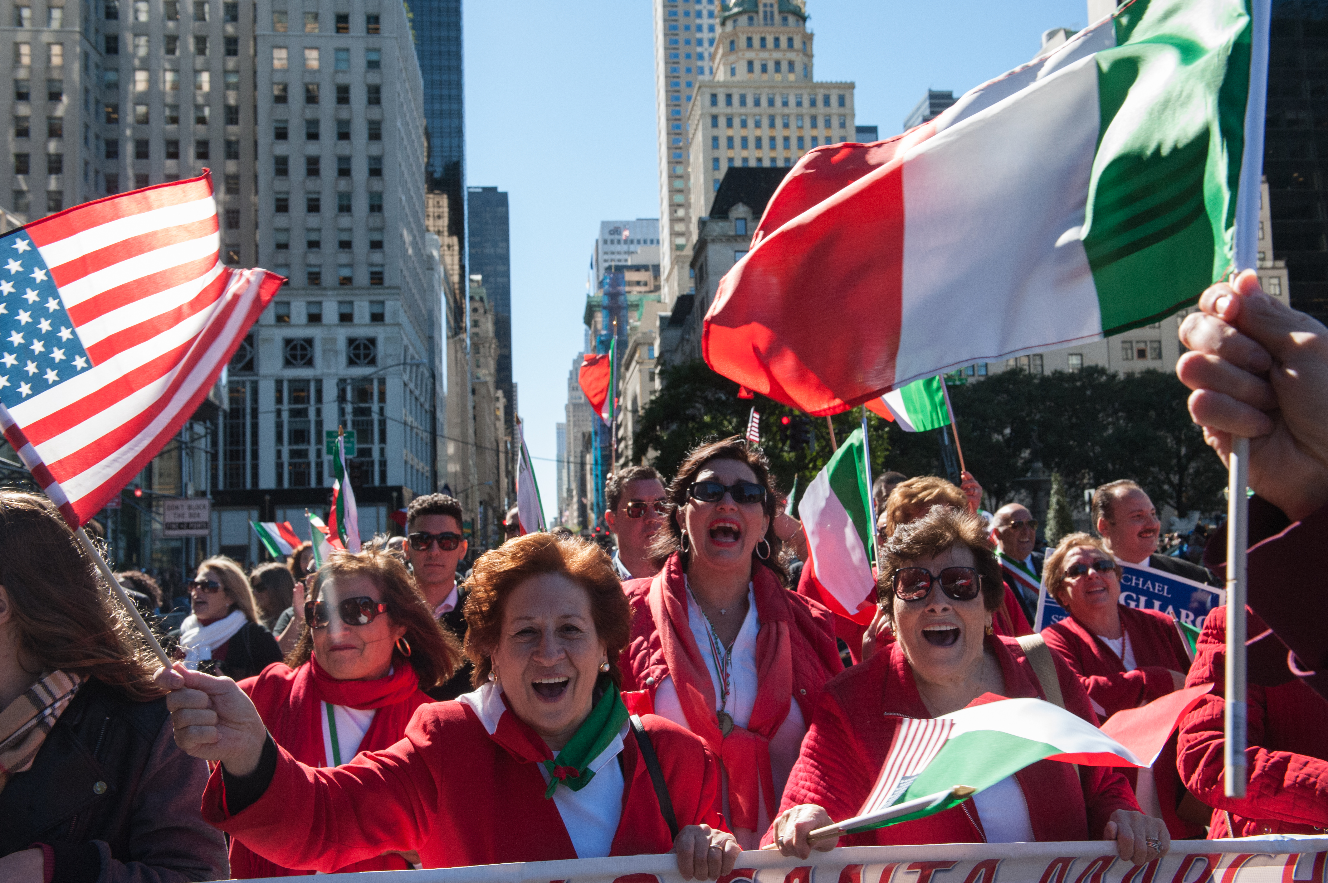 Parade goers at the annual Columbus Day Parade in New York City. People are wearing red jackets and holding up the flags of Italy and the United States. The people are in the street and there are various city buildings in the background.
