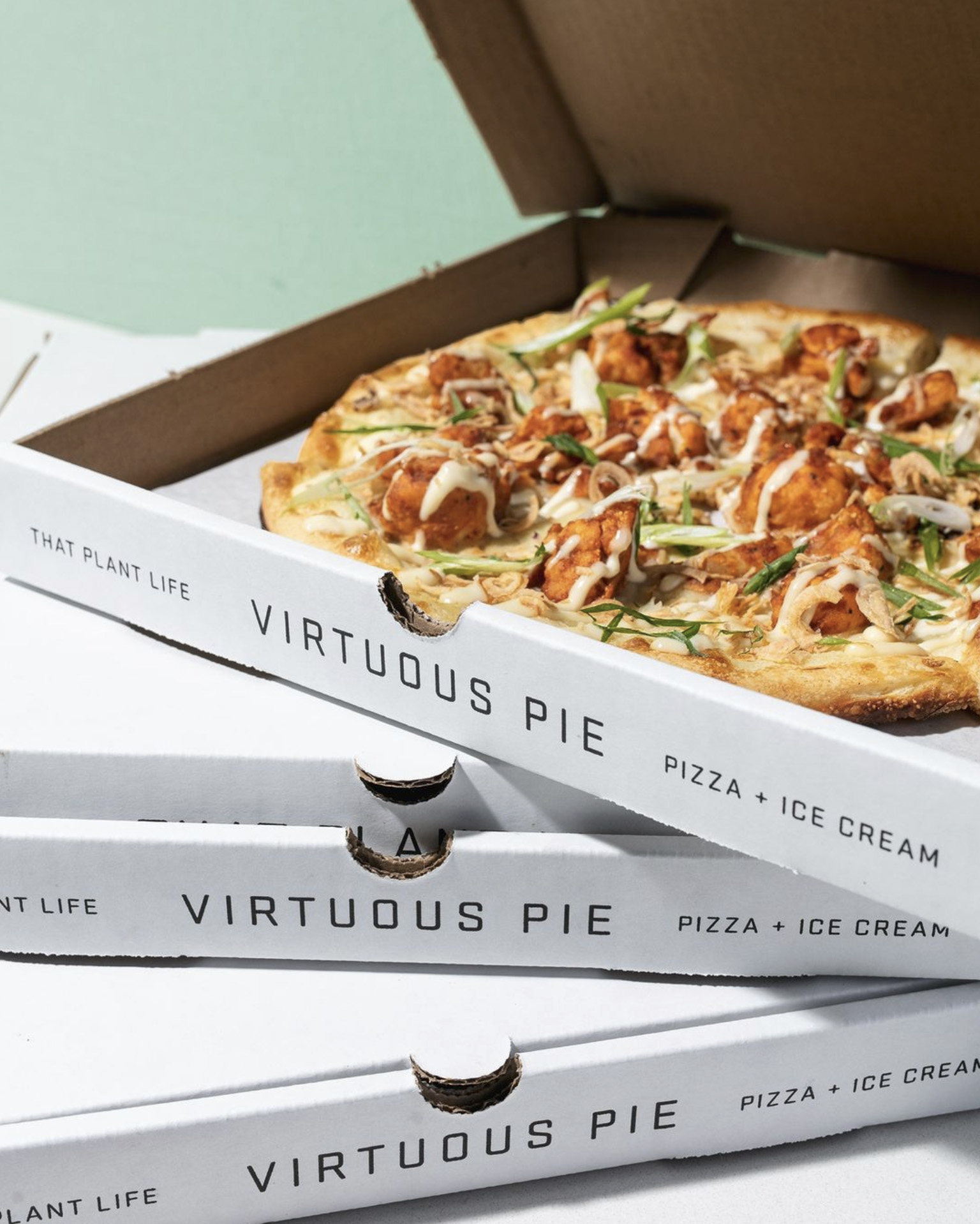 A photo of the Virtuous Pie Stranger Wings vegan buffalo cauliflower pie in a pizza box stacked on top of more pizza boxes against a light green background.