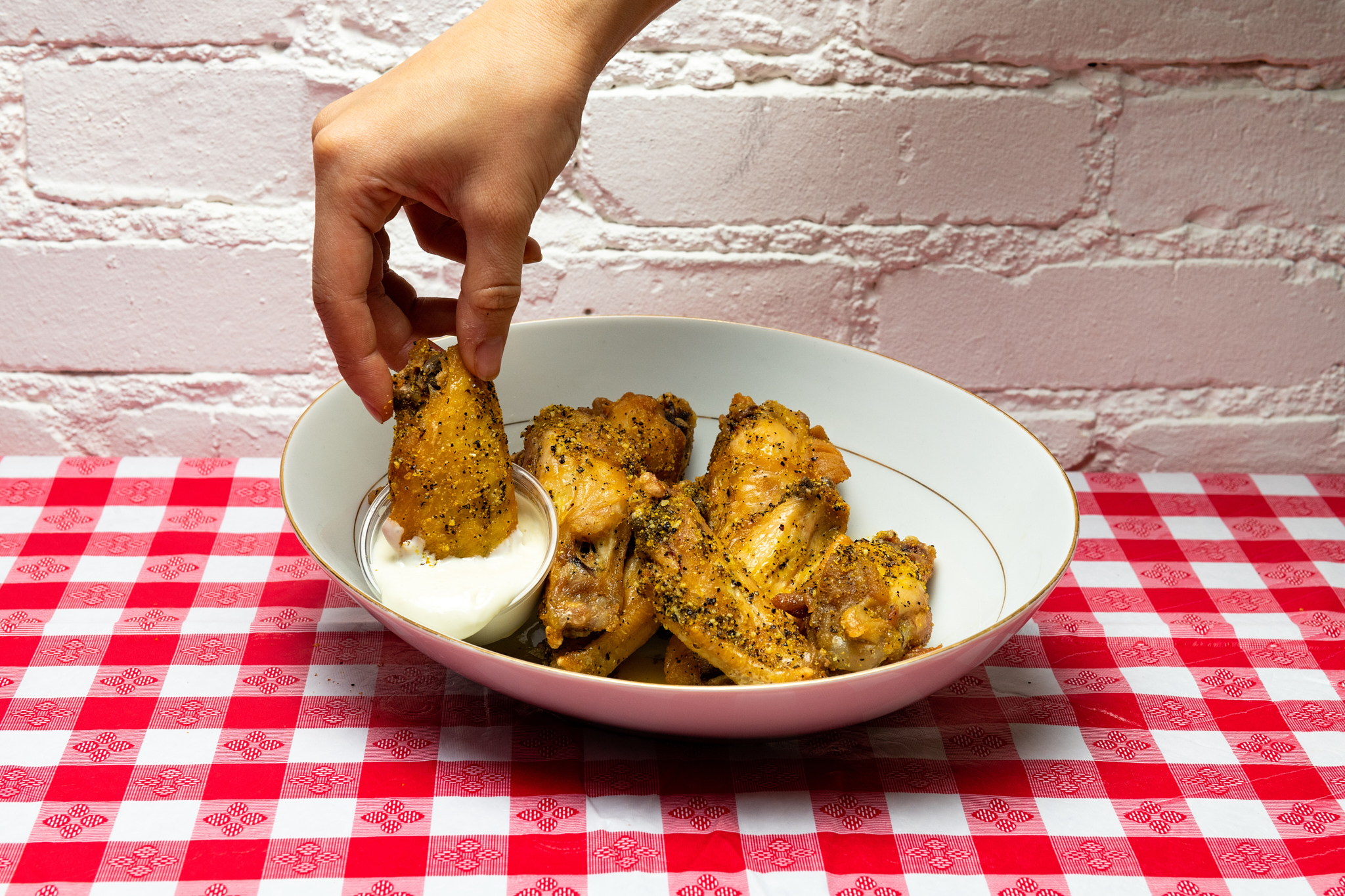 A hand grabs a chicken wing coated in spices from a bowl and dips it into a white sauce.