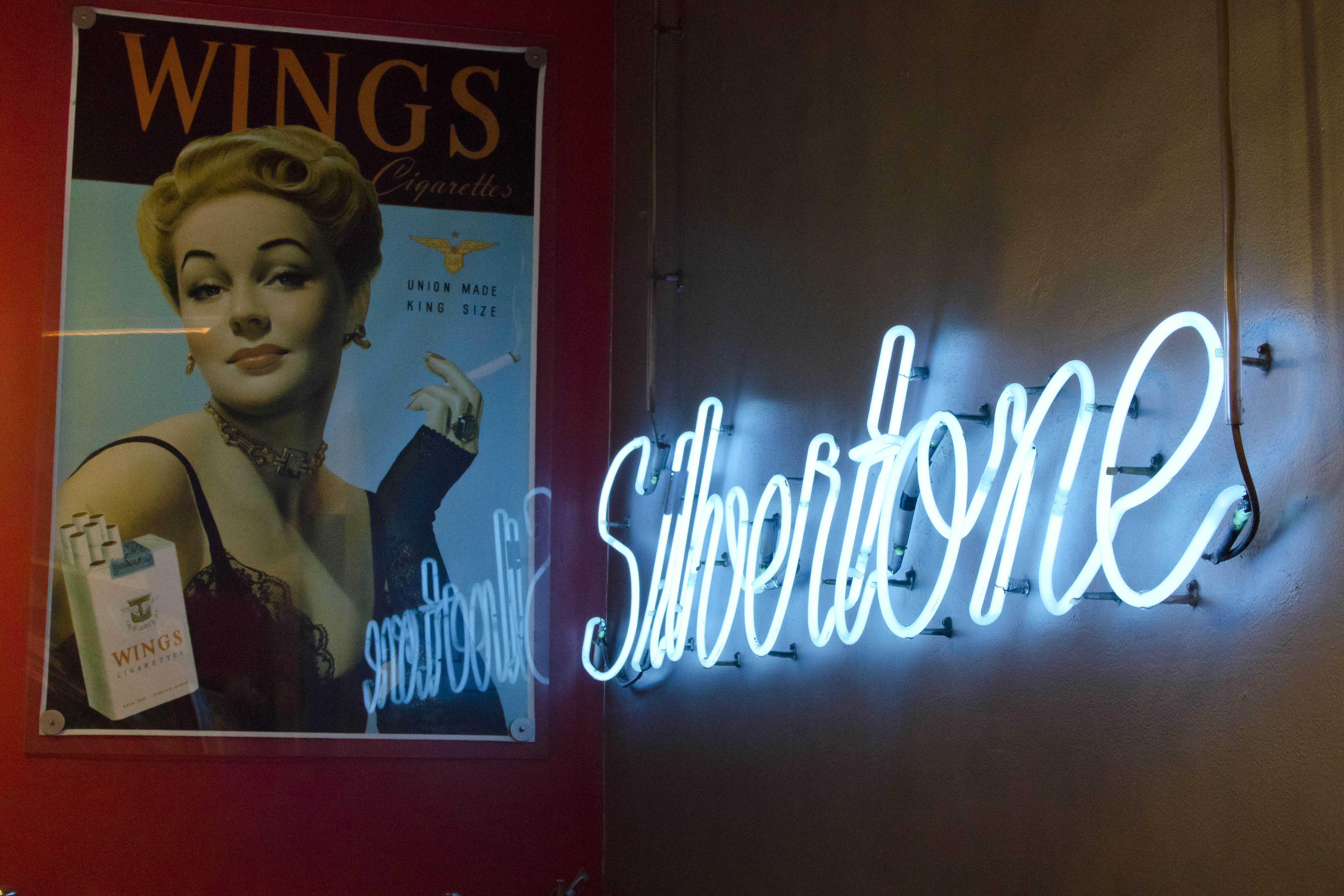A vintage cigarette poster hangs on a red wall, across a corner from a blue neon sign that reads Silvertone.