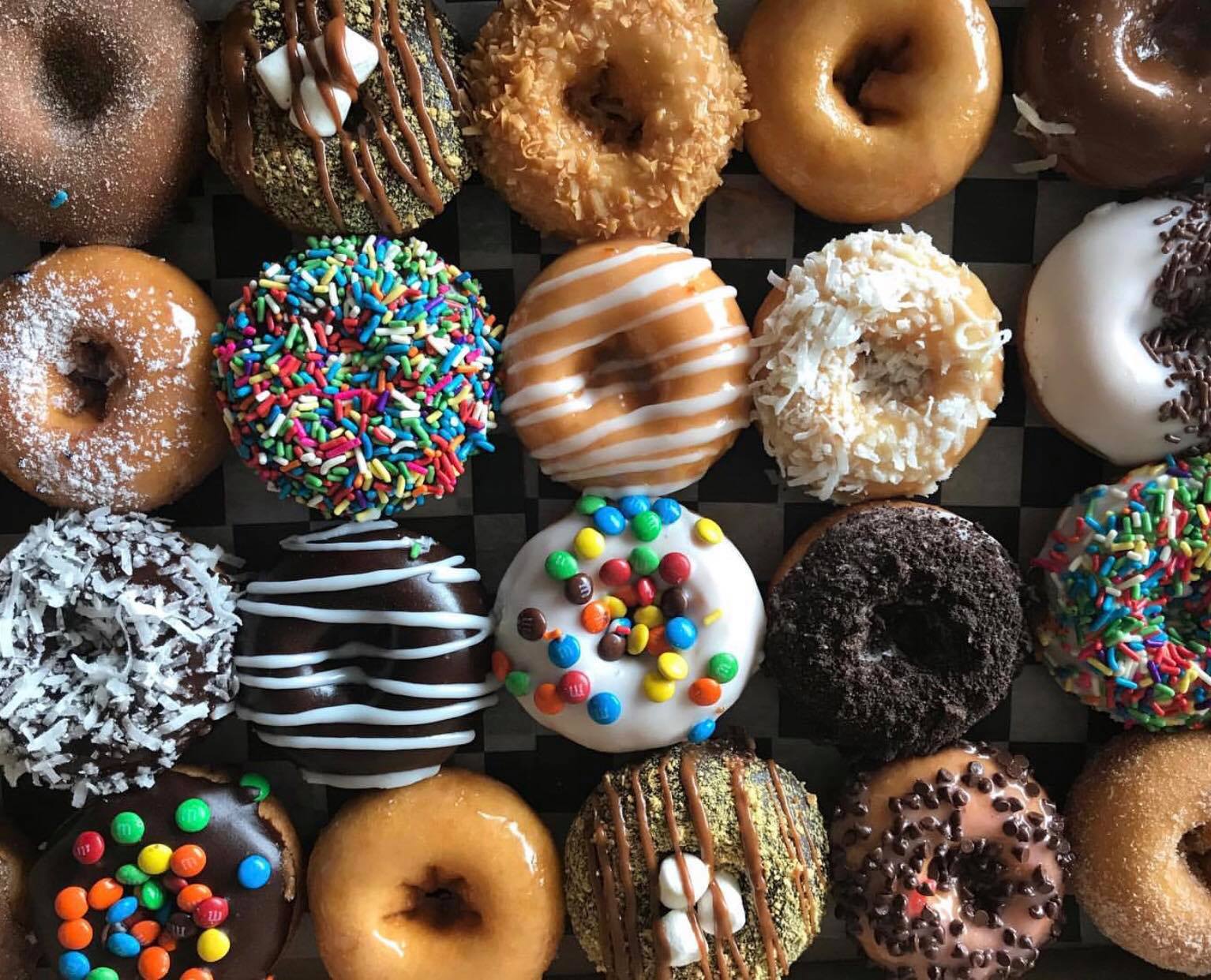A large assortment of doughnuts including doughnuts topped with sprinkles, chocolate glaze, and coconut.