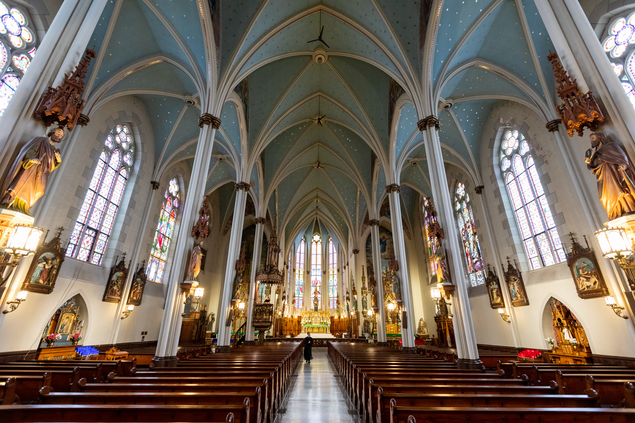 The nave of a church with rows of pews and a center aisle. The ceiling has many vaults and curves and is painted light blue. There’s also arched windows on either side, some of which have stained glass.