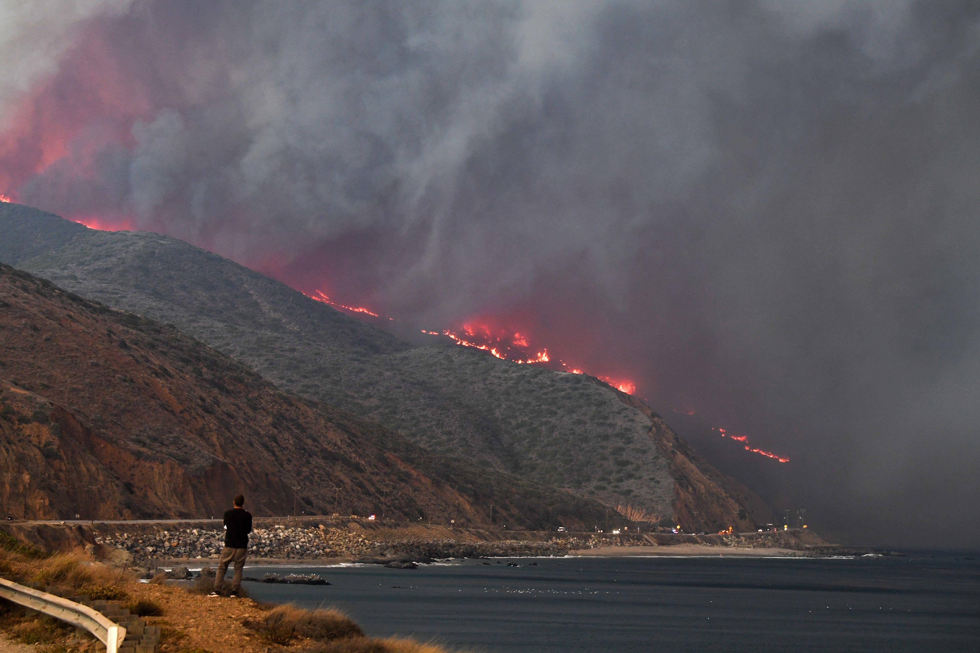 A photo of a man standing on the beach, looking back at nearby hillsides on fire.
