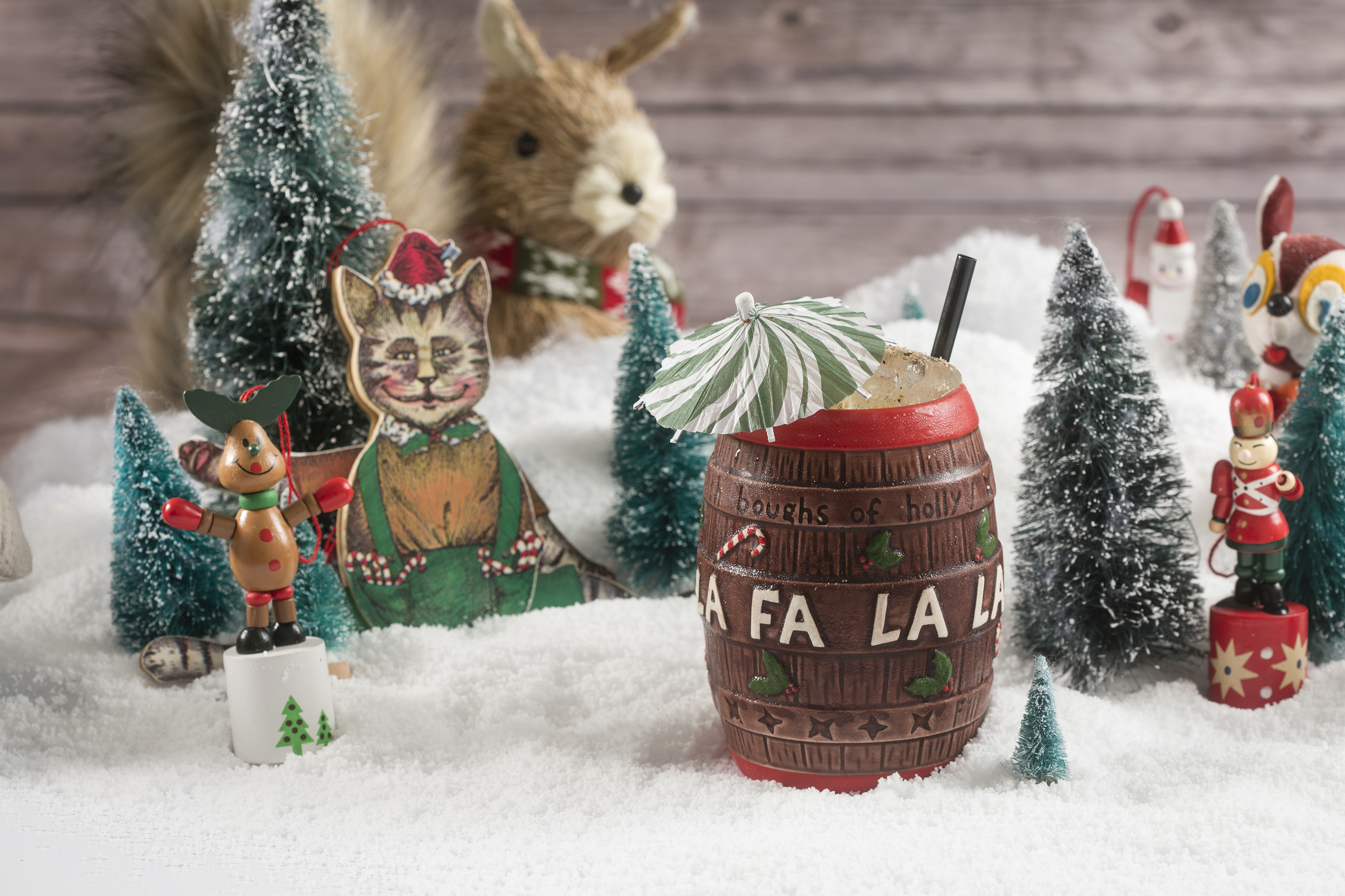 A tiki drink is served in a Christmas-themed cup shaped like a barrel with “fa la la” written on it. The drink is set on a Christmas backdrop with fake snow, trees, and other festive knick-knacks.