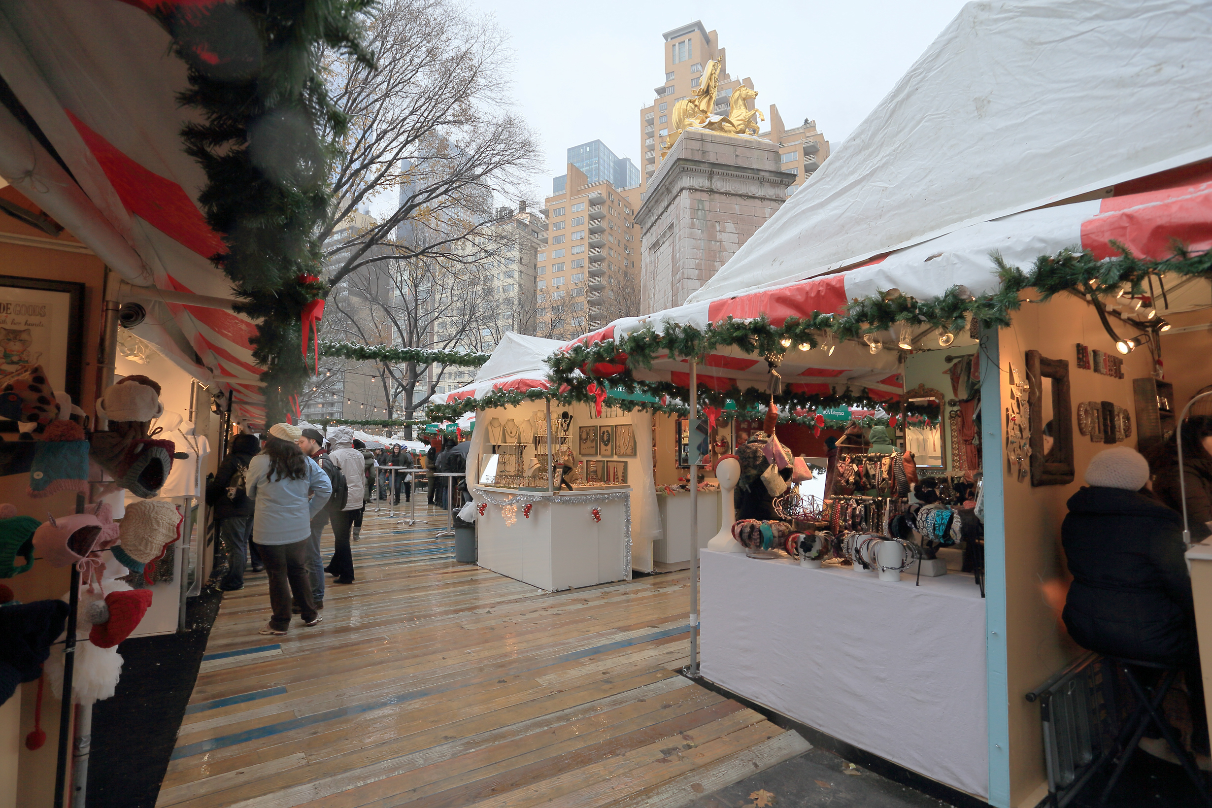 A holiday market in New York City. There are various market stalls with wares. Each stall is decorated with garlands. There are tall city buildings in the background.