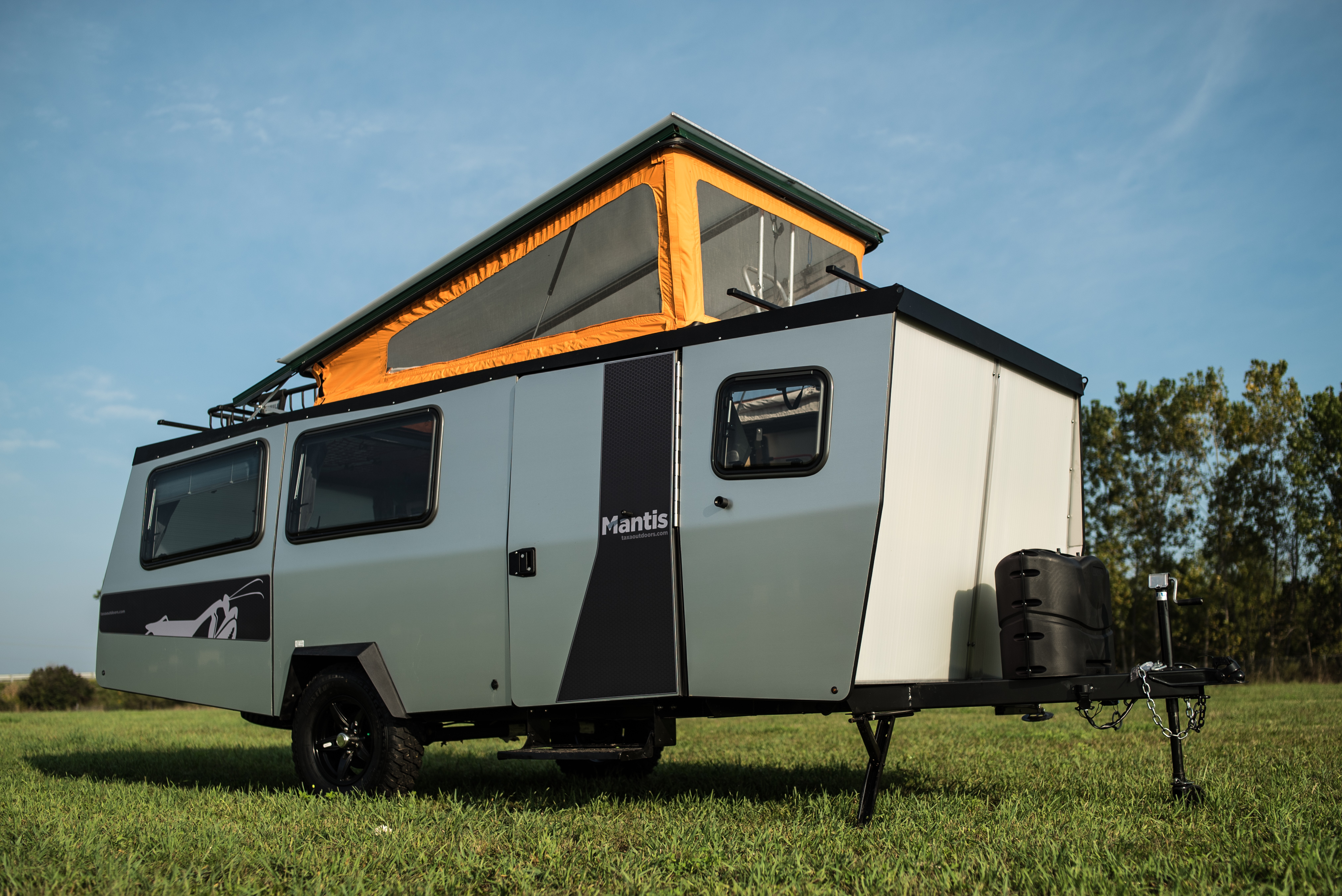 A white and tan camper with an inflatable orange tent on top of the camper. The camper is parked in a grassy field.