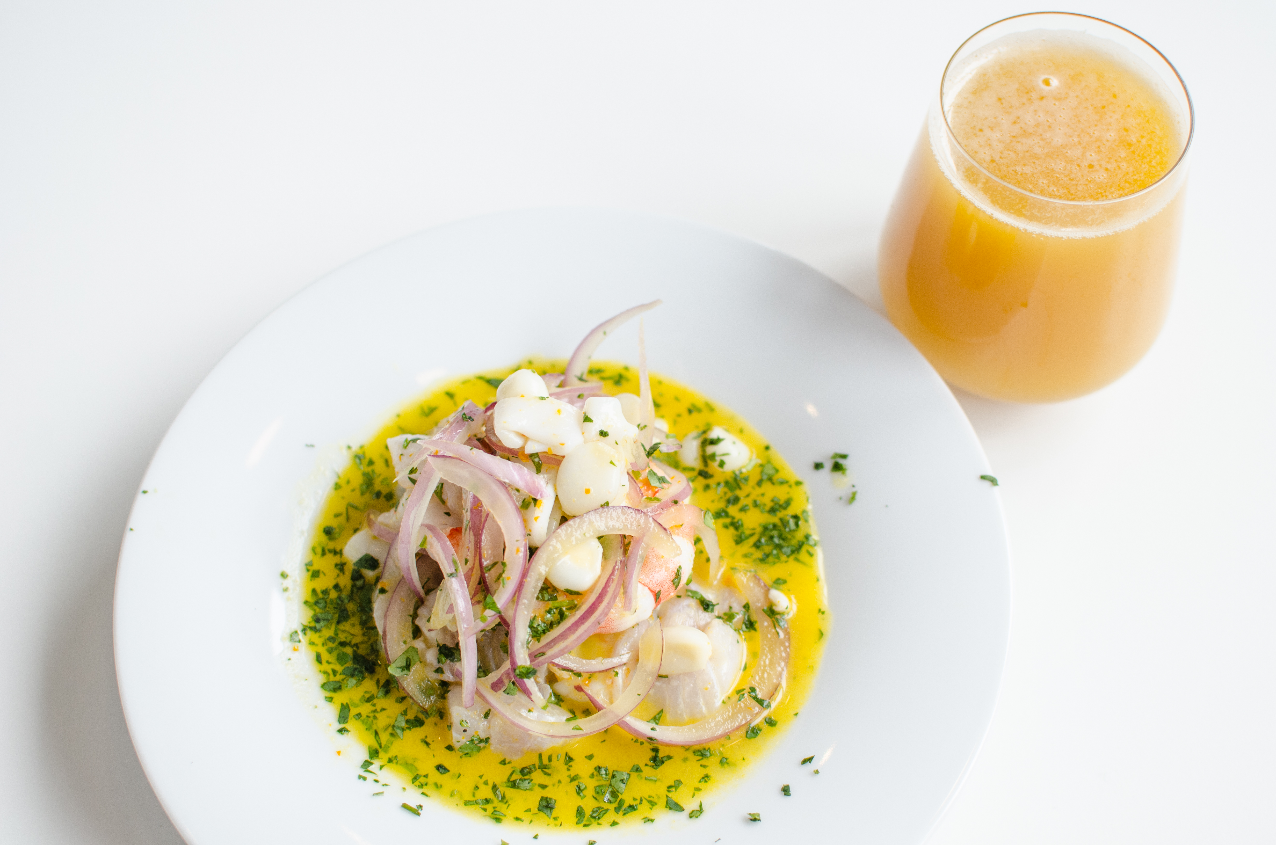 A vibrantly colored portion of ceviche is presented on a white plate on a white table, with a glass of beer on the side.