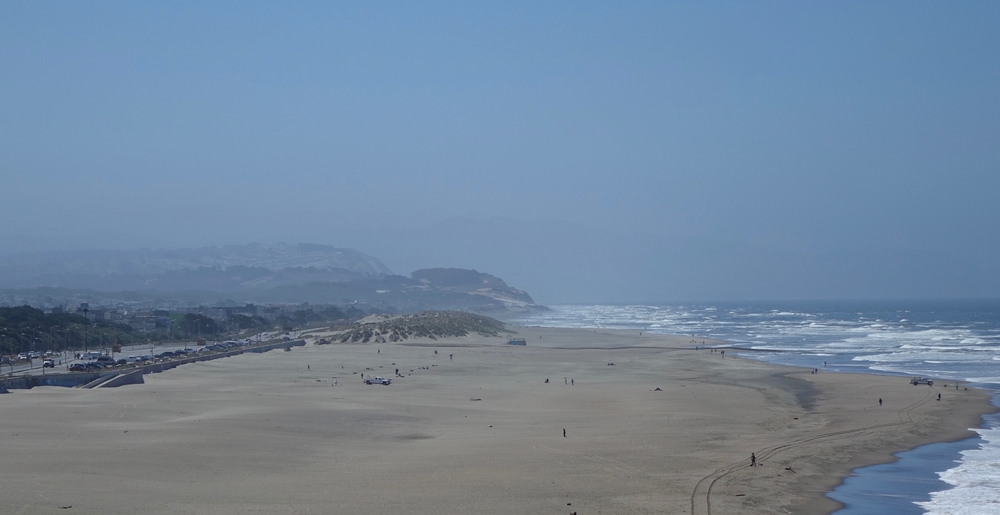 A photo of Ocean Beach with fog rolling in, with the Great Highway visible in the distance.