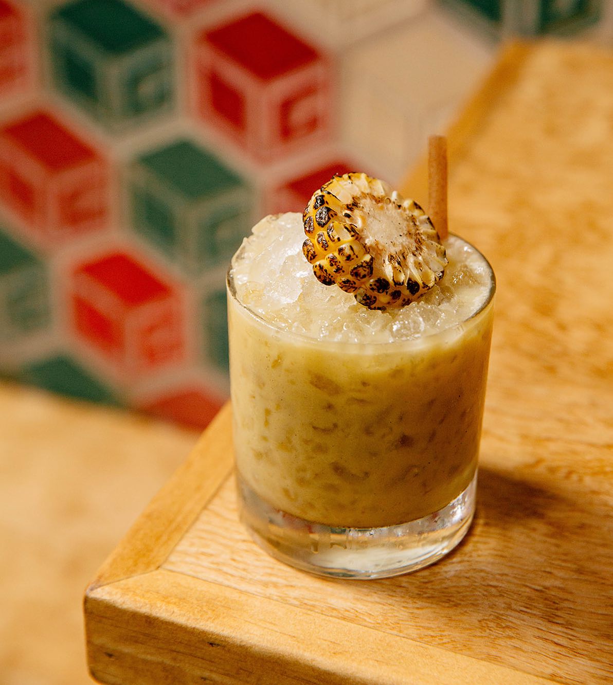 A tumbler filled with crushed ice, a beige liquid, and garnished with a charred wheel of corn and a straw. The drink is sitting on a wooden table.