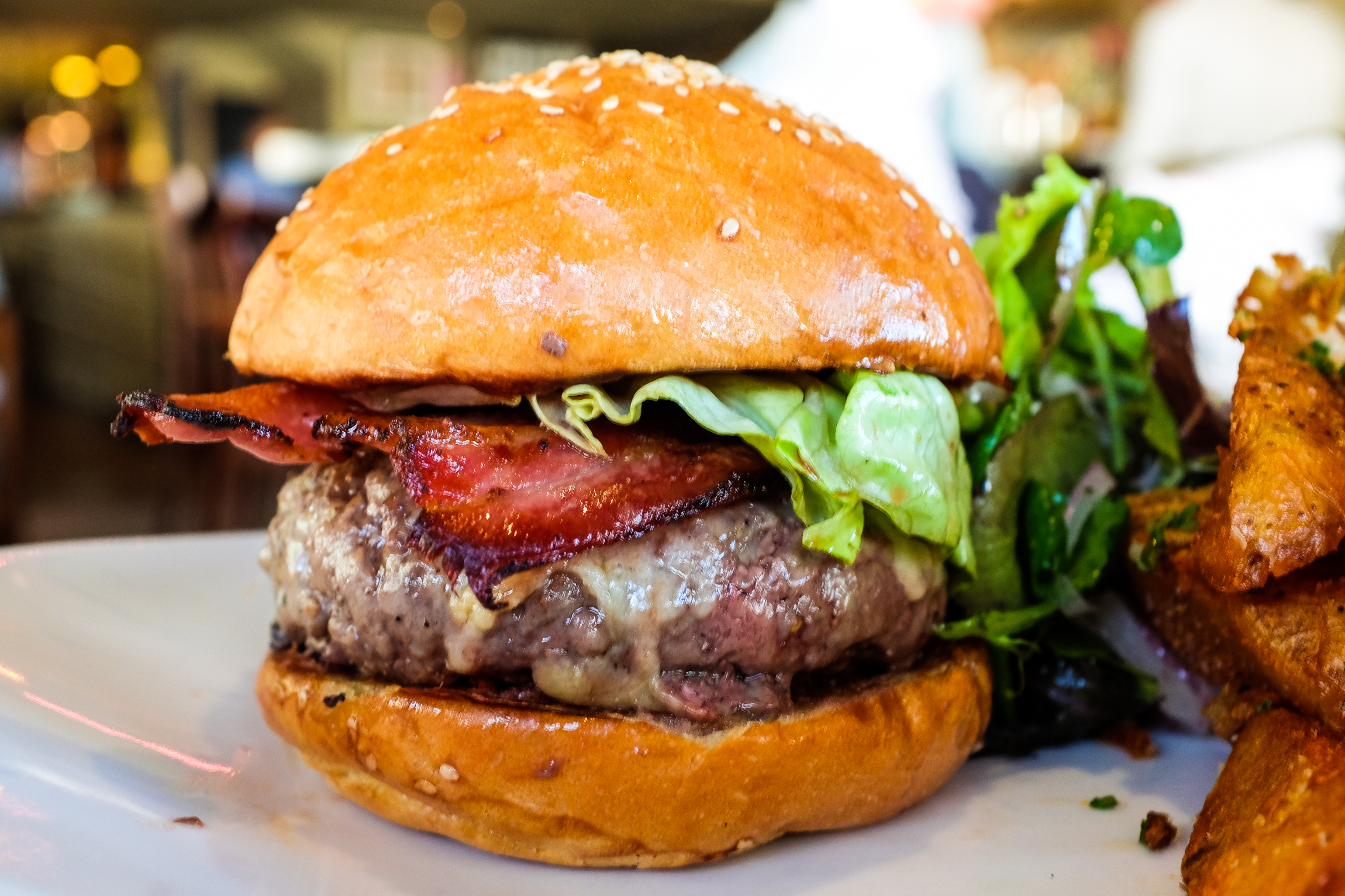Closeup on a burger featuring a thick patty, bacon, lettuce, and cheese, with thick fries on the edge of the frame