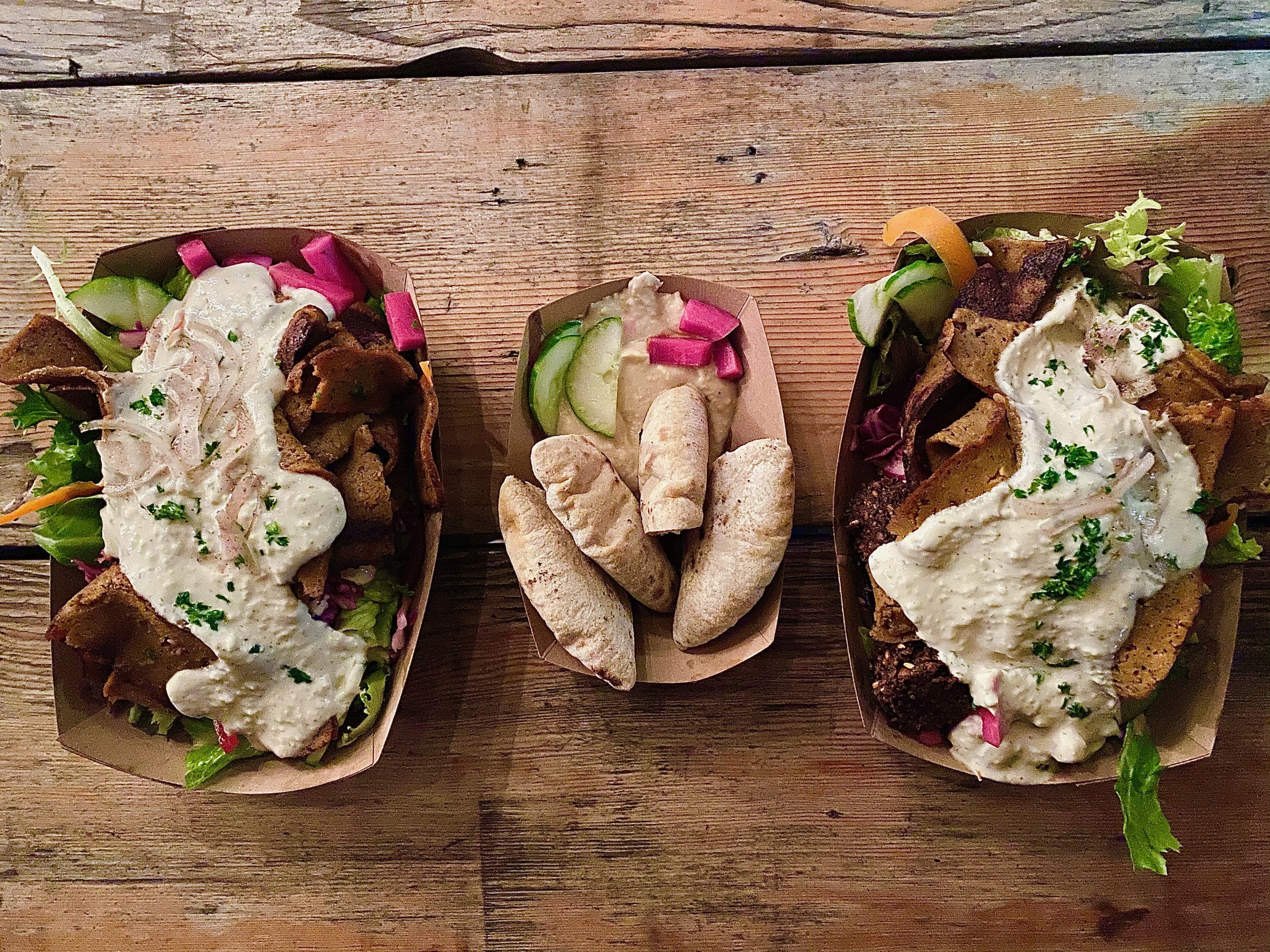 Three boats full of hummus, pita, and vegetables sit on a wood table