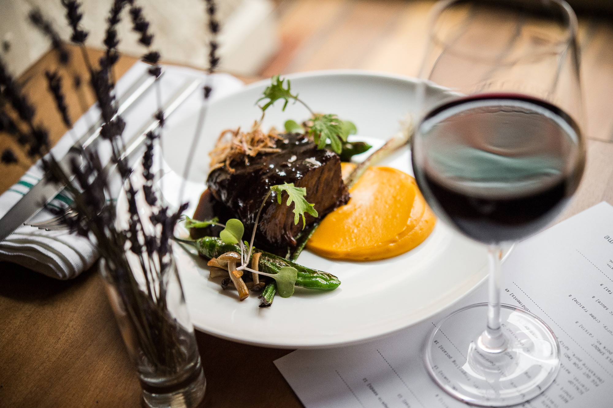A glass of red wine and flowers in front of a white plate with a saucy meat dish at Tuome.