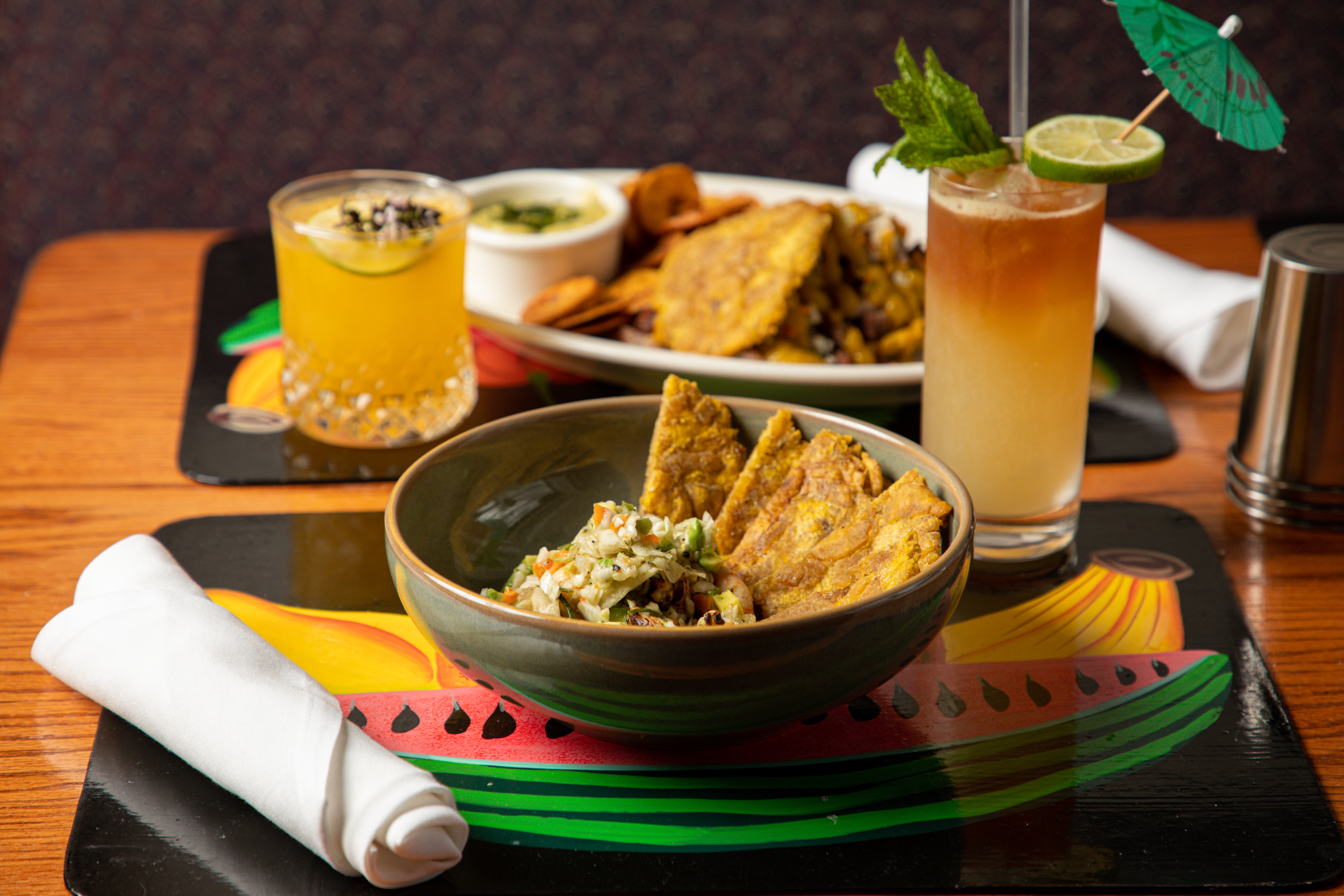 A table setting with two bright colored cocktails and plates of food; one with grilled shrimp pikliz and plantains and one with a plantain pulled pork sandwich platter.