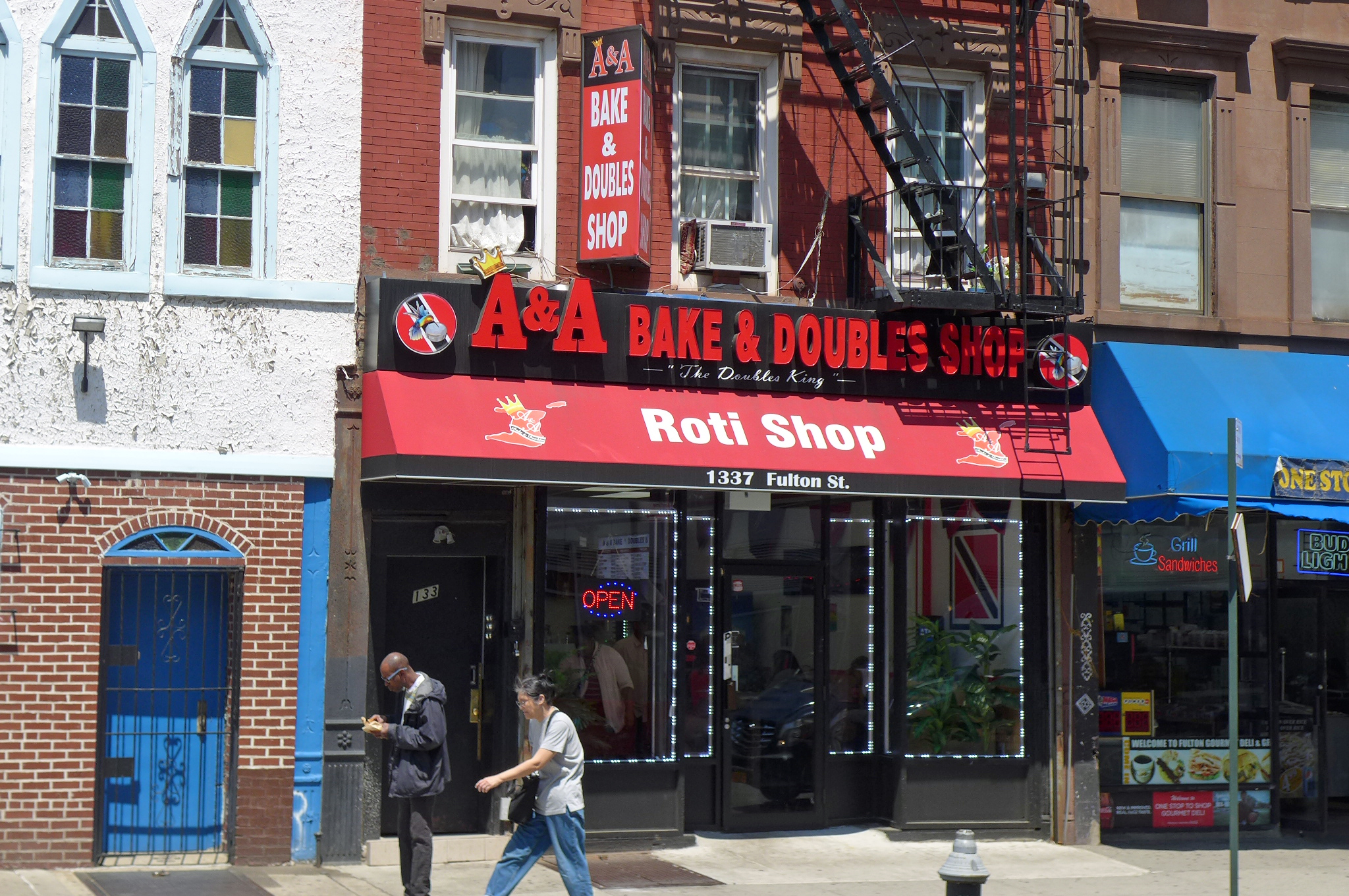 Two people walk in front a red storefront whose neon sign reads “A&amp;A Bake &amp; Doubles Roti Shop”
