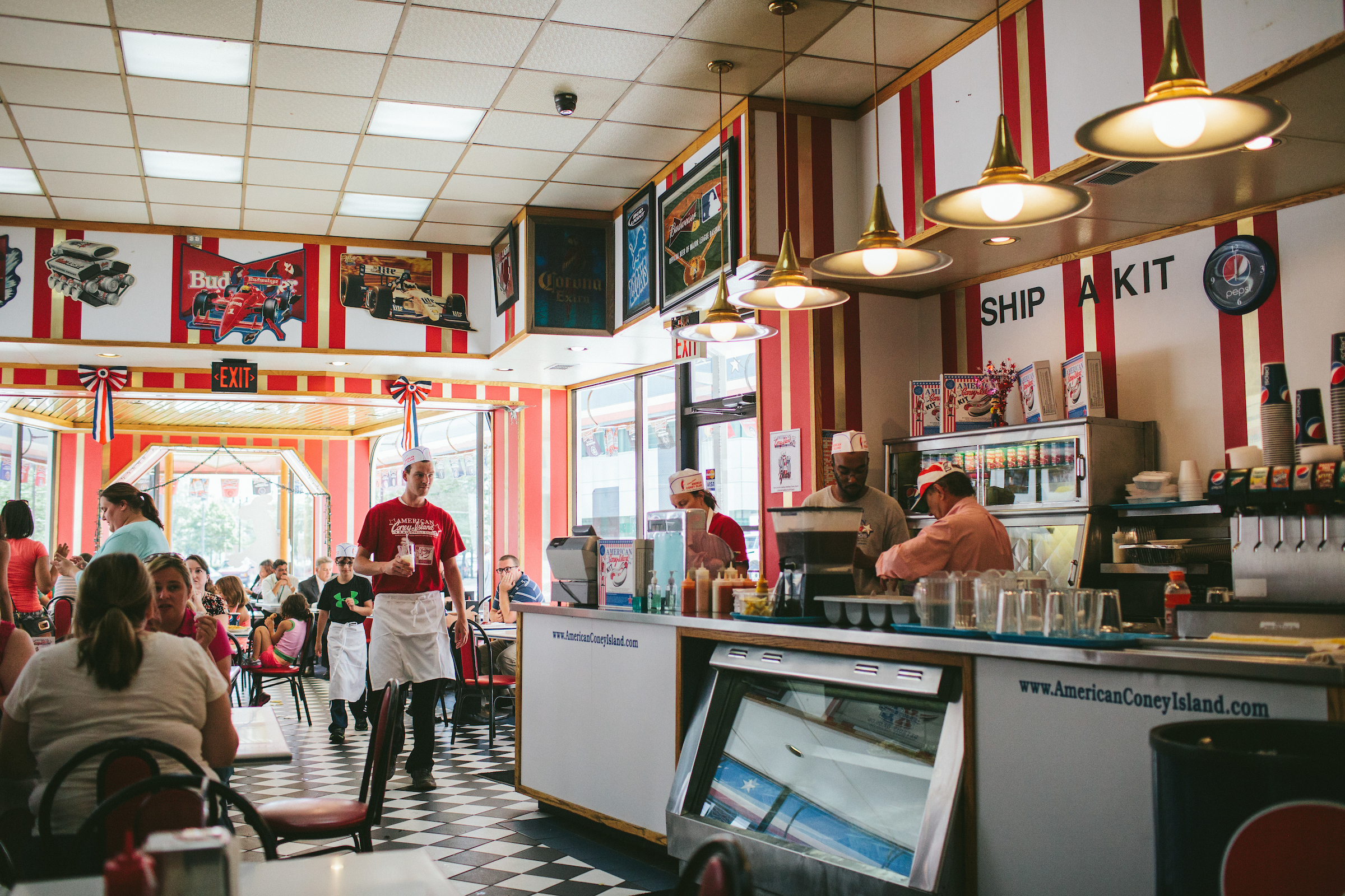 A server in a red T-shirt and apron walking through a full, retro style dining room at American Coney Island.