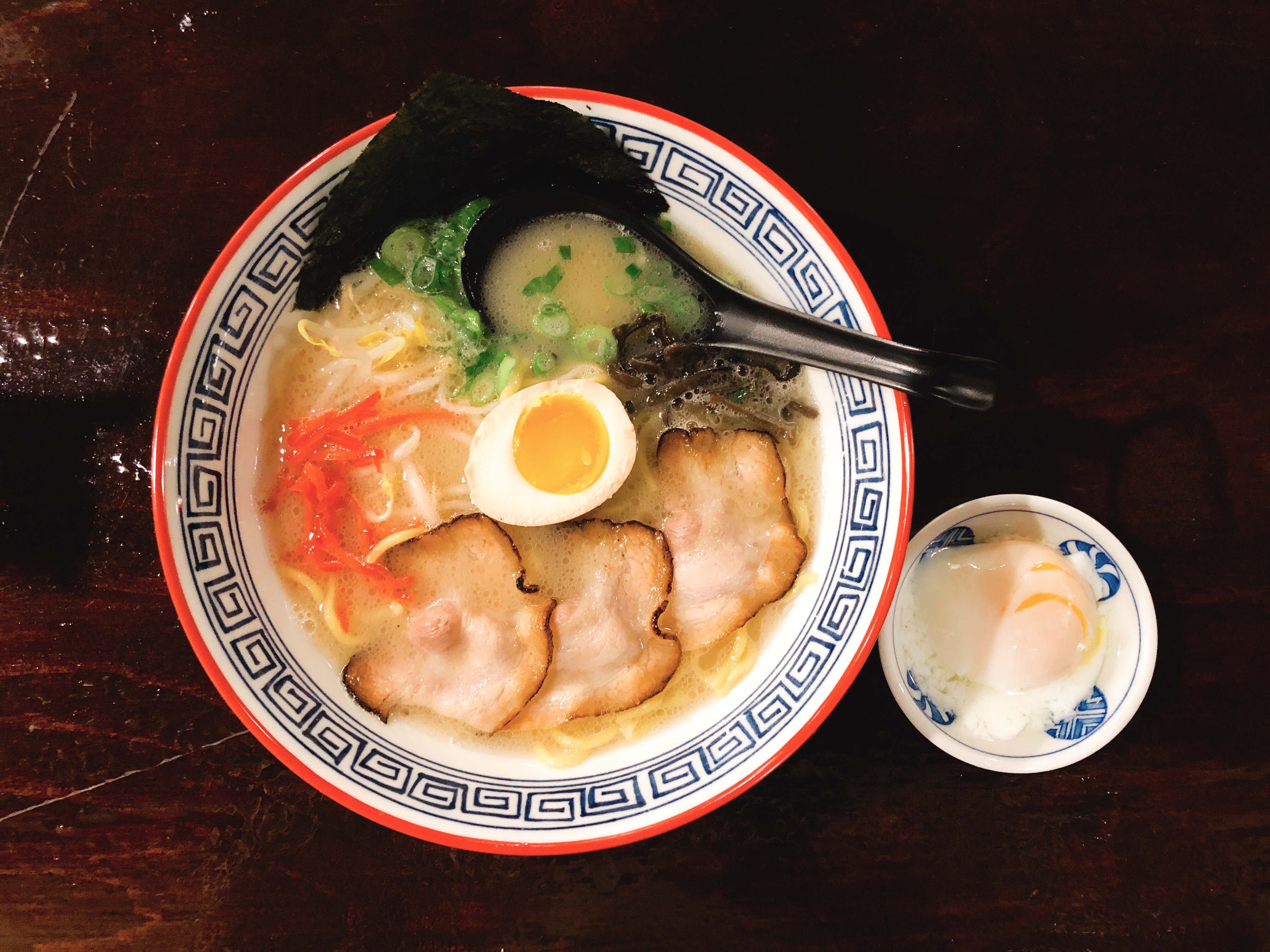 A bowl of ramen with a black spoon, slices of meat, chopped vegetables, and a soft egg.