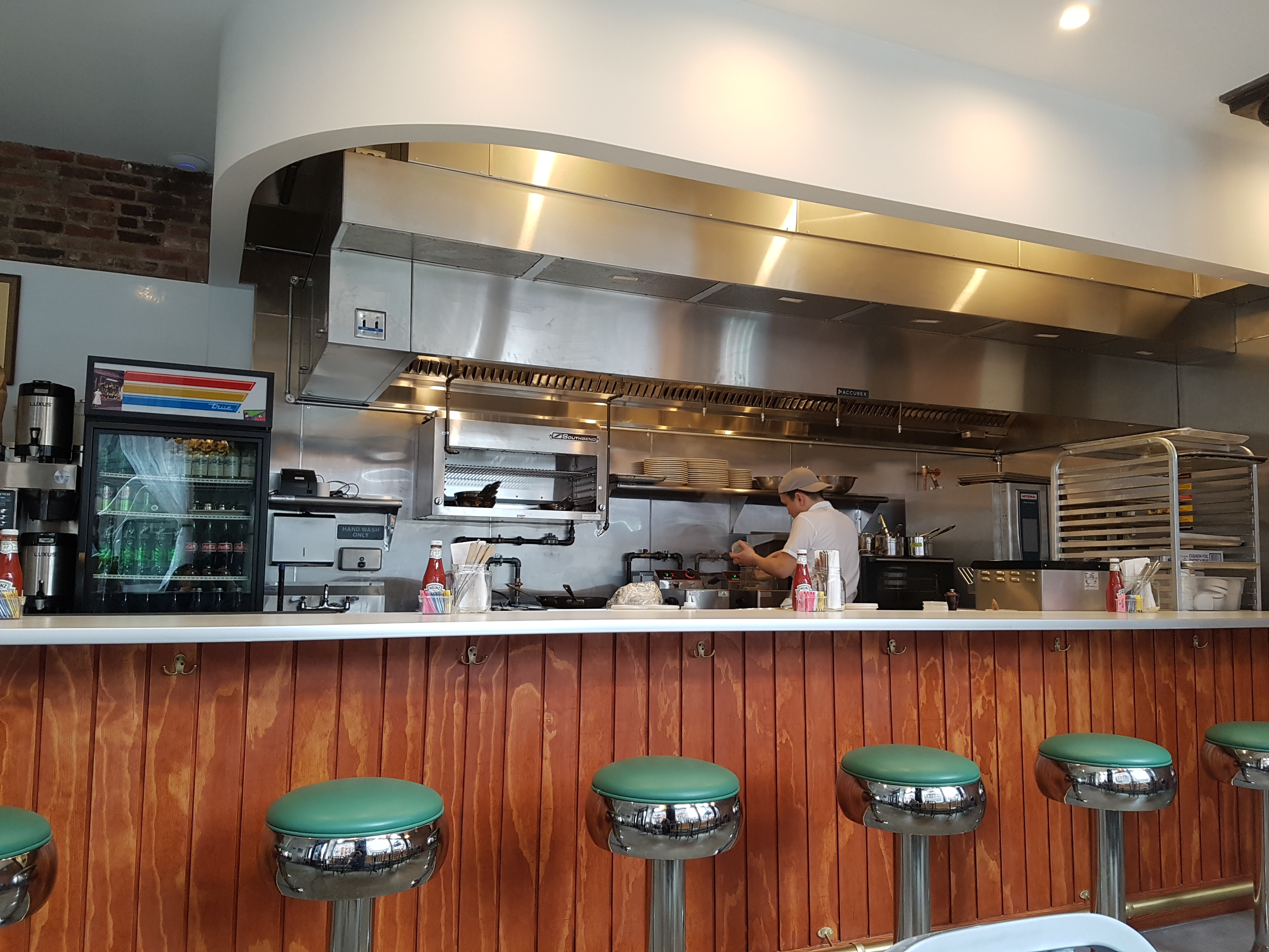 Green bar stools are located in front of a wooden diner counter, and the kitchen can be seen in the background