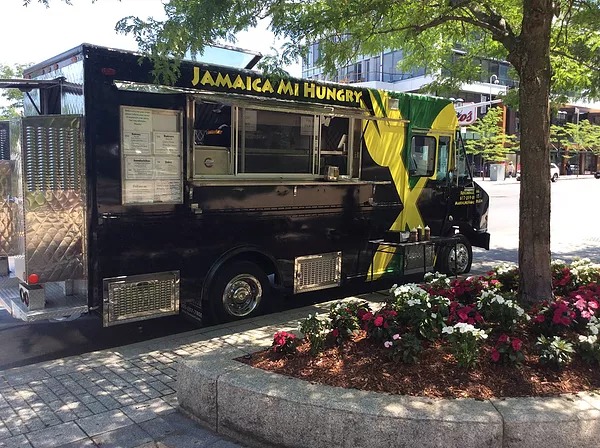 A black food truck painted with yellow and green parks on the street in front of a sidewalk planter with flowers and a tree