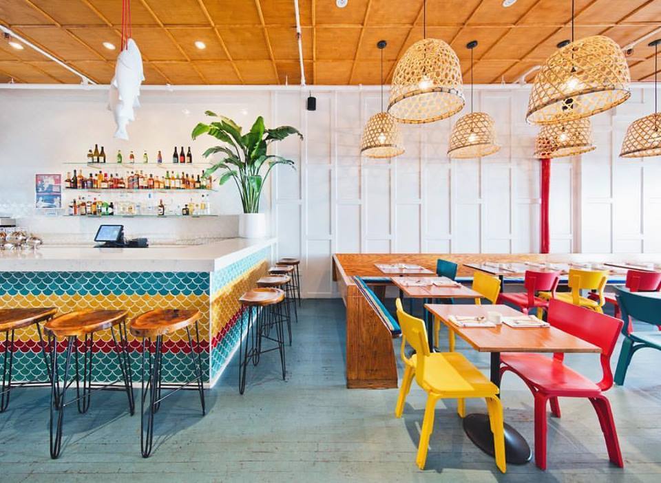 A colorful restaurant interior with a light wooden ceiling, wooden tables, and red, yellow, and blue chairs.