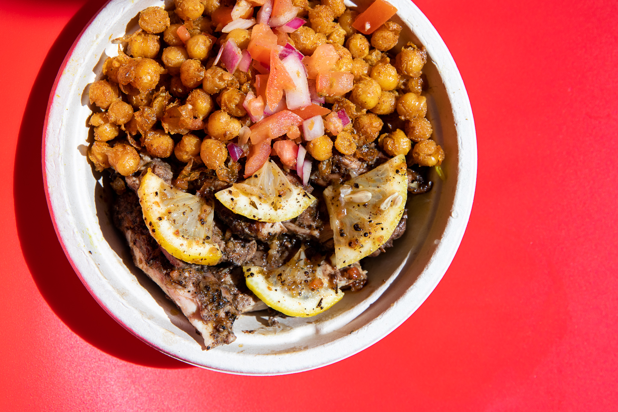 A bowl filled with half fried chickpeas and chopped red onion and half season chicken with quarters of thin-sliced lemon on a red background. 