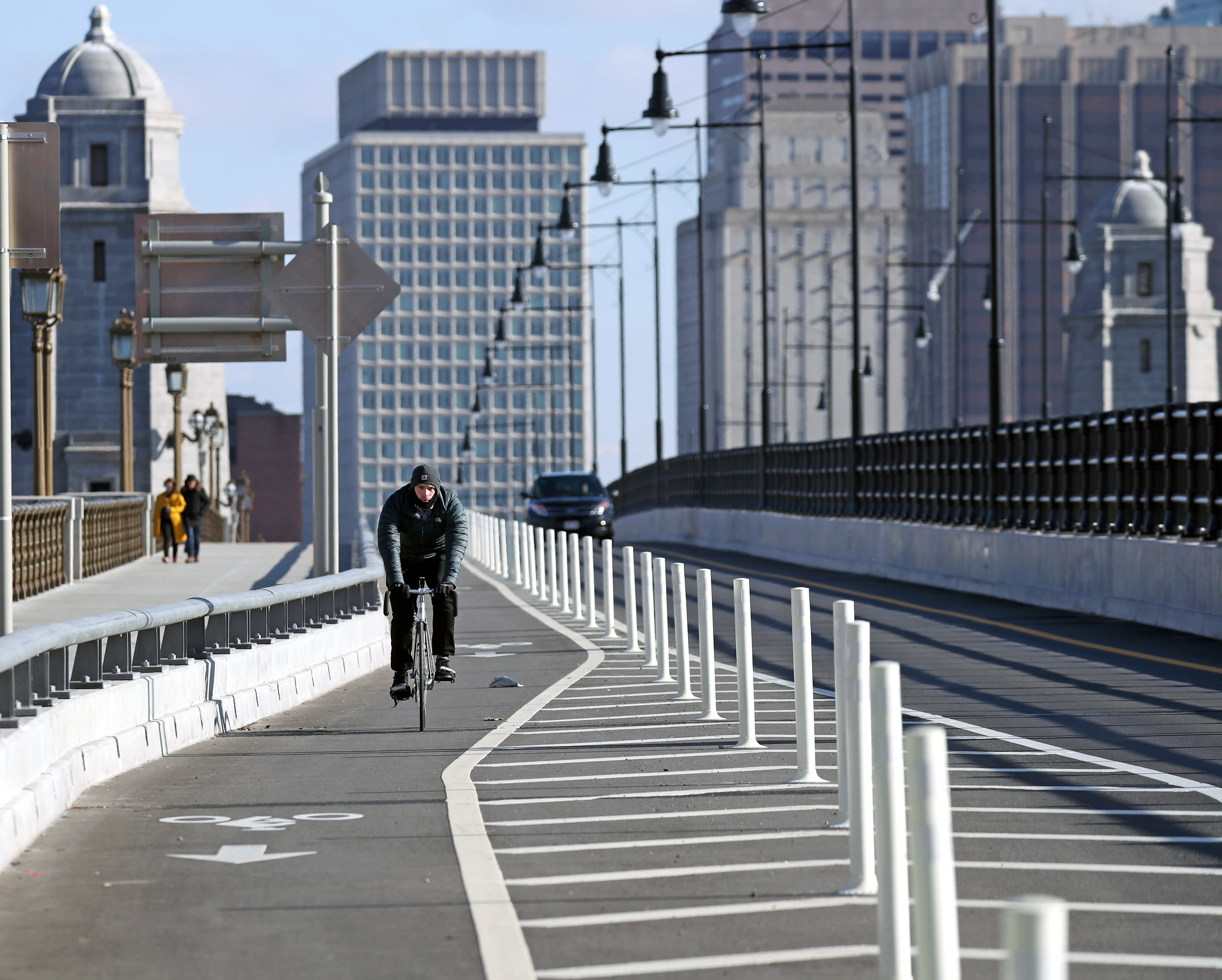 A lone bicyclist riding in a protected bike lane on a bridge and there are buildings in the background.