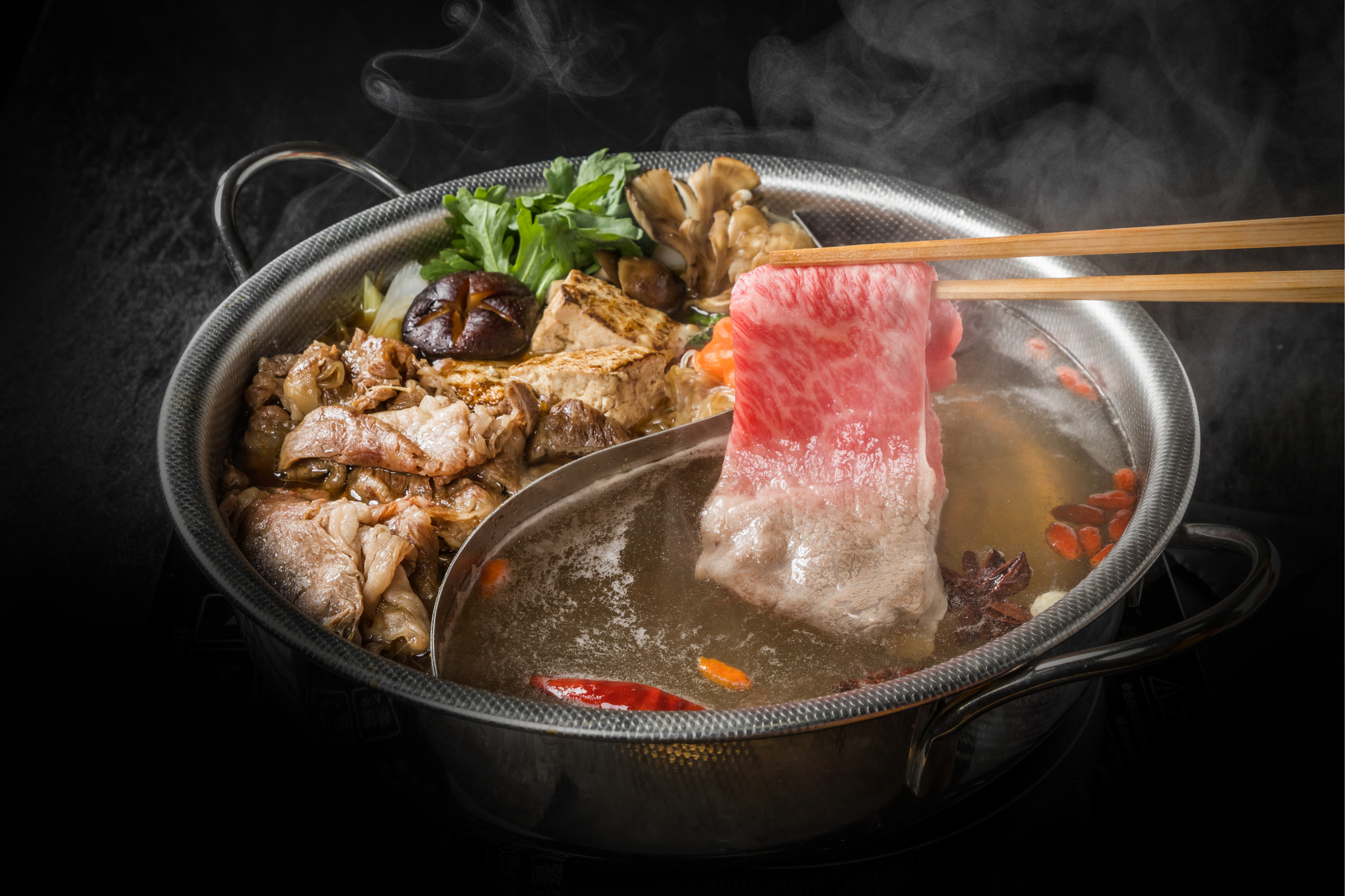 A pair of chopsticks holding a piece of meat over a bowl of Chinese hot pot soup