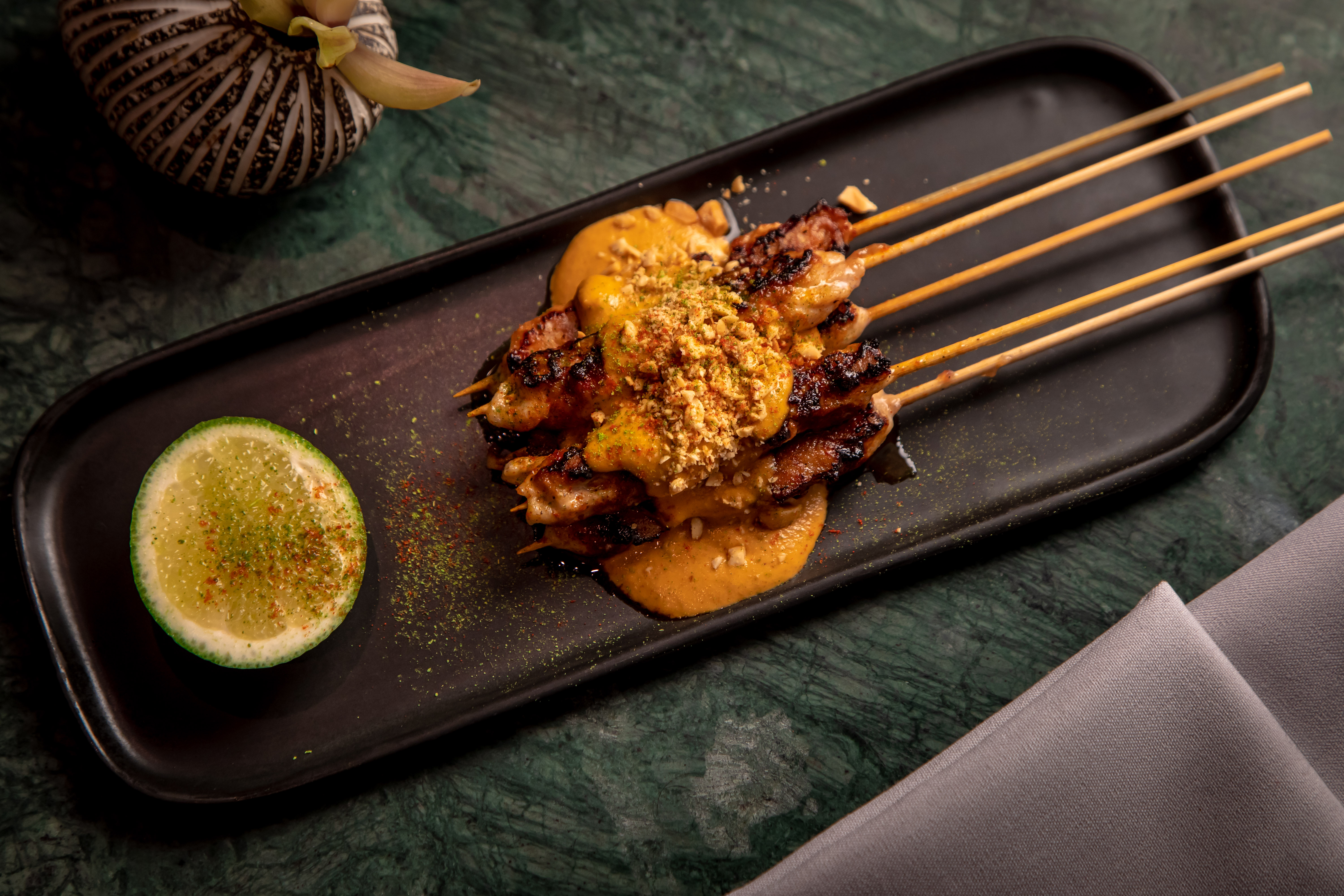 Chicken satays with peanut sauce laid on a black, oblong platter with a halved lime on the side.