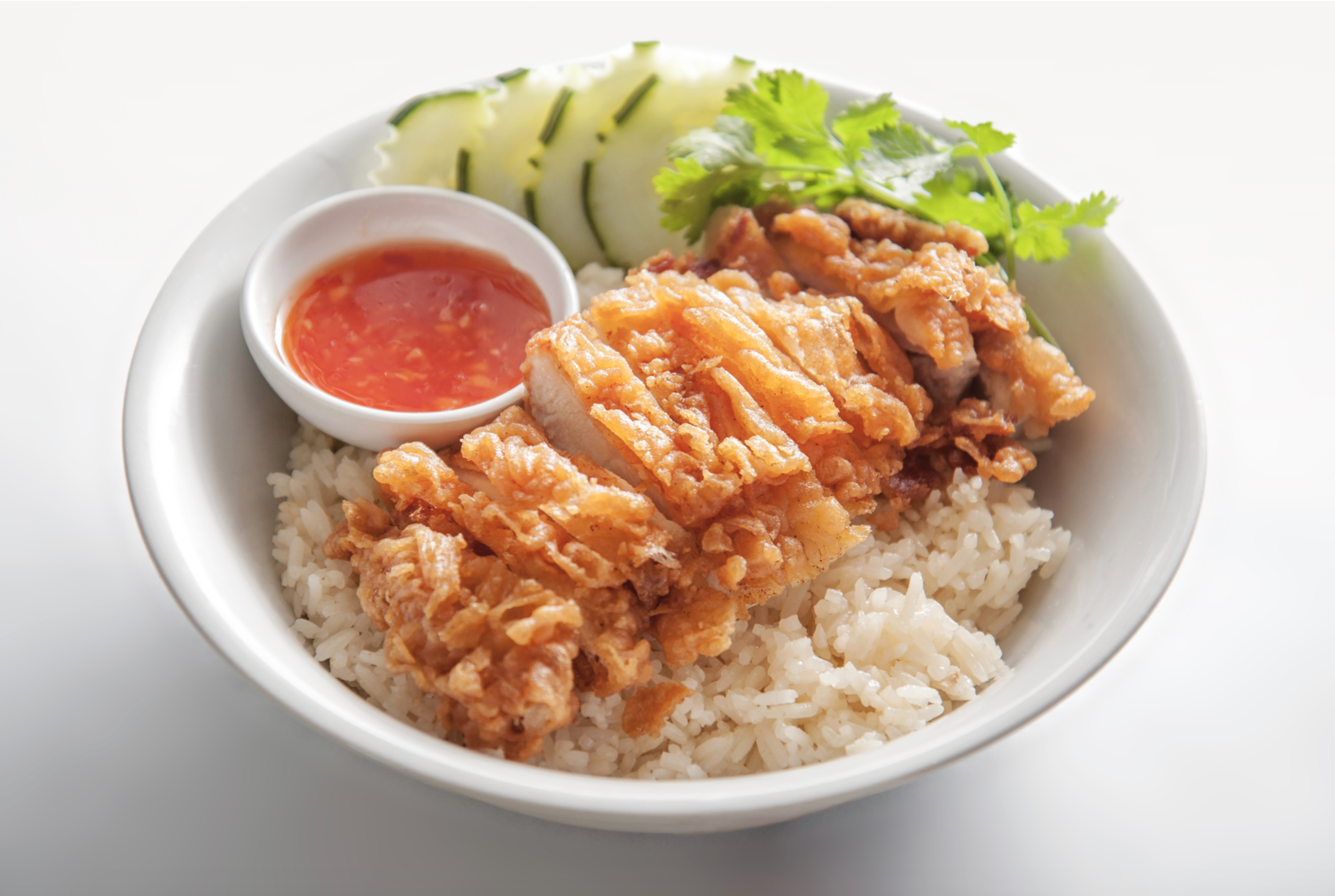 A bowl of light-colored fried chicken rests atop rice next to cucumber, greens, and a side of red chili sauce