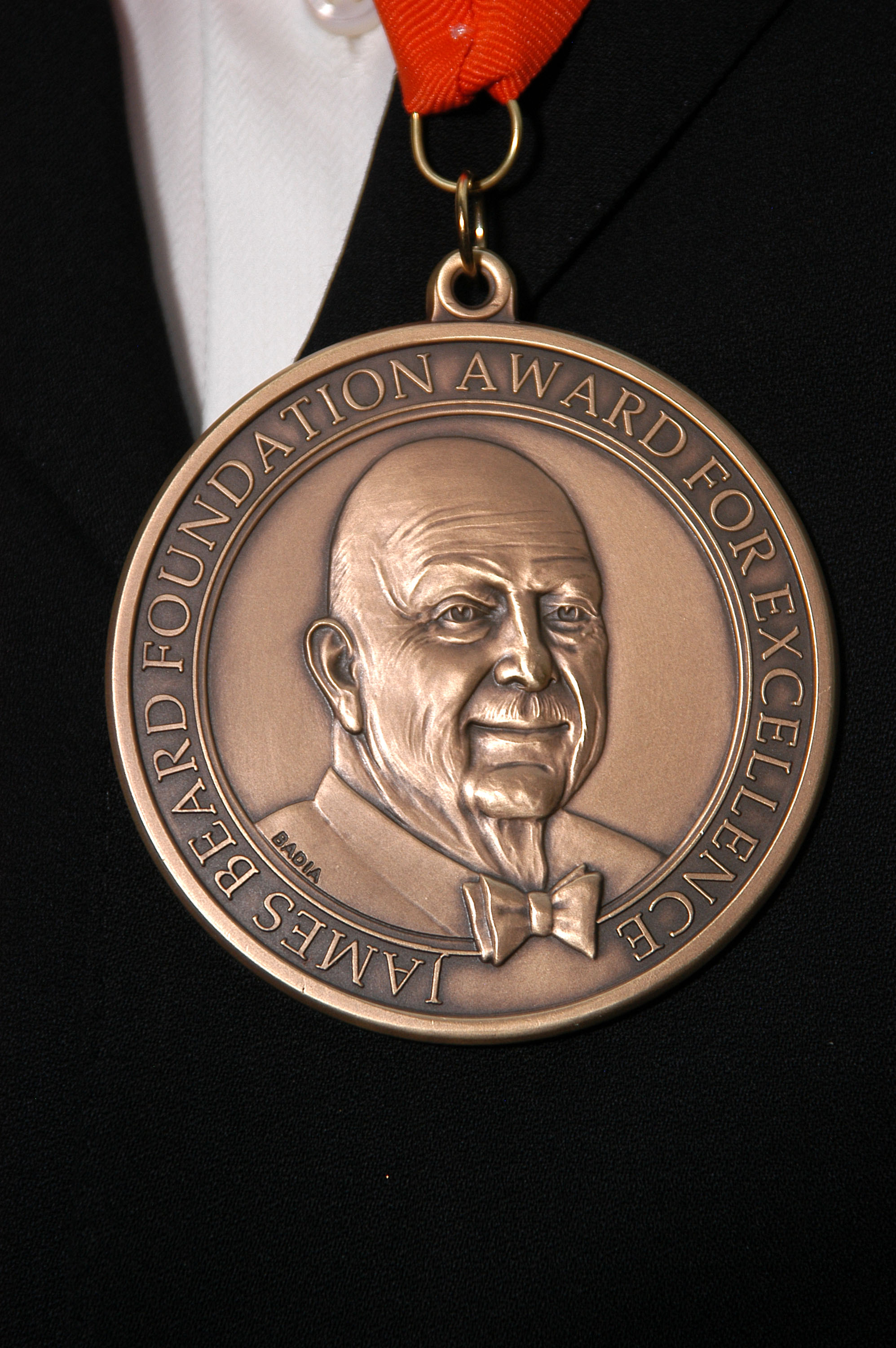 The James Beard award medal, cast in bronze, with the likeness of James Beard surrounded with the words “James Beard Foundation Award for Excellence” against a black background. 