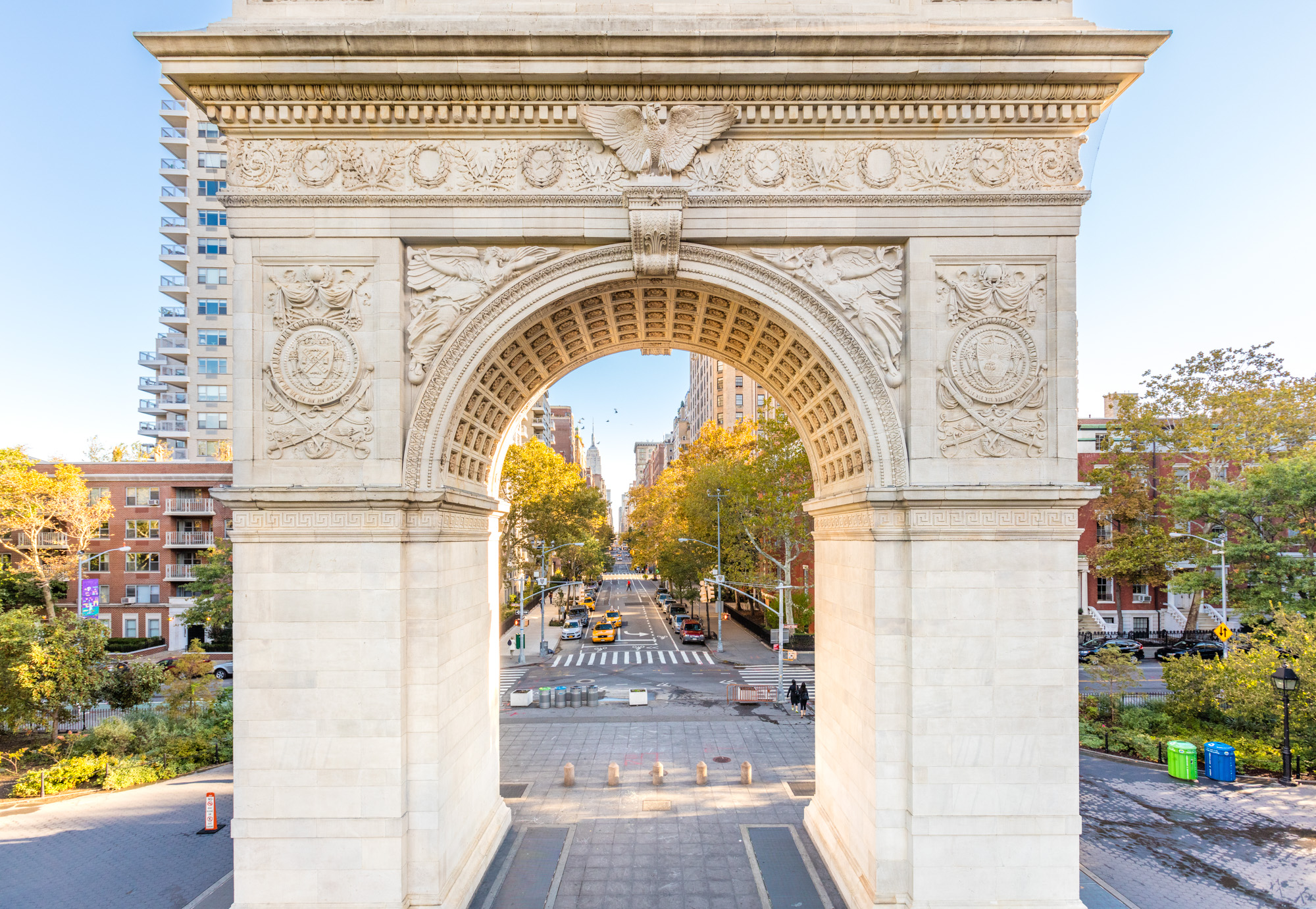 A large white arch in a New York City Park, with cars driving up a street visible through the archway. 