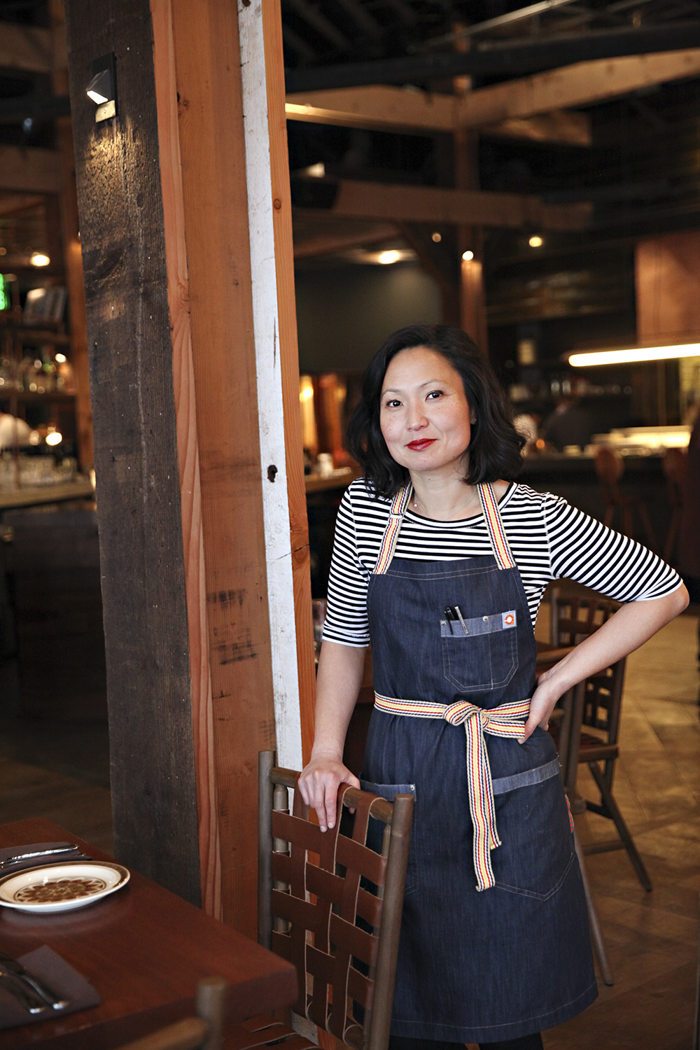 Ann Kim wearing a black and white striped shirt and blue apron leans on a chair, one hand on her hip inside the dining room at Young Joni