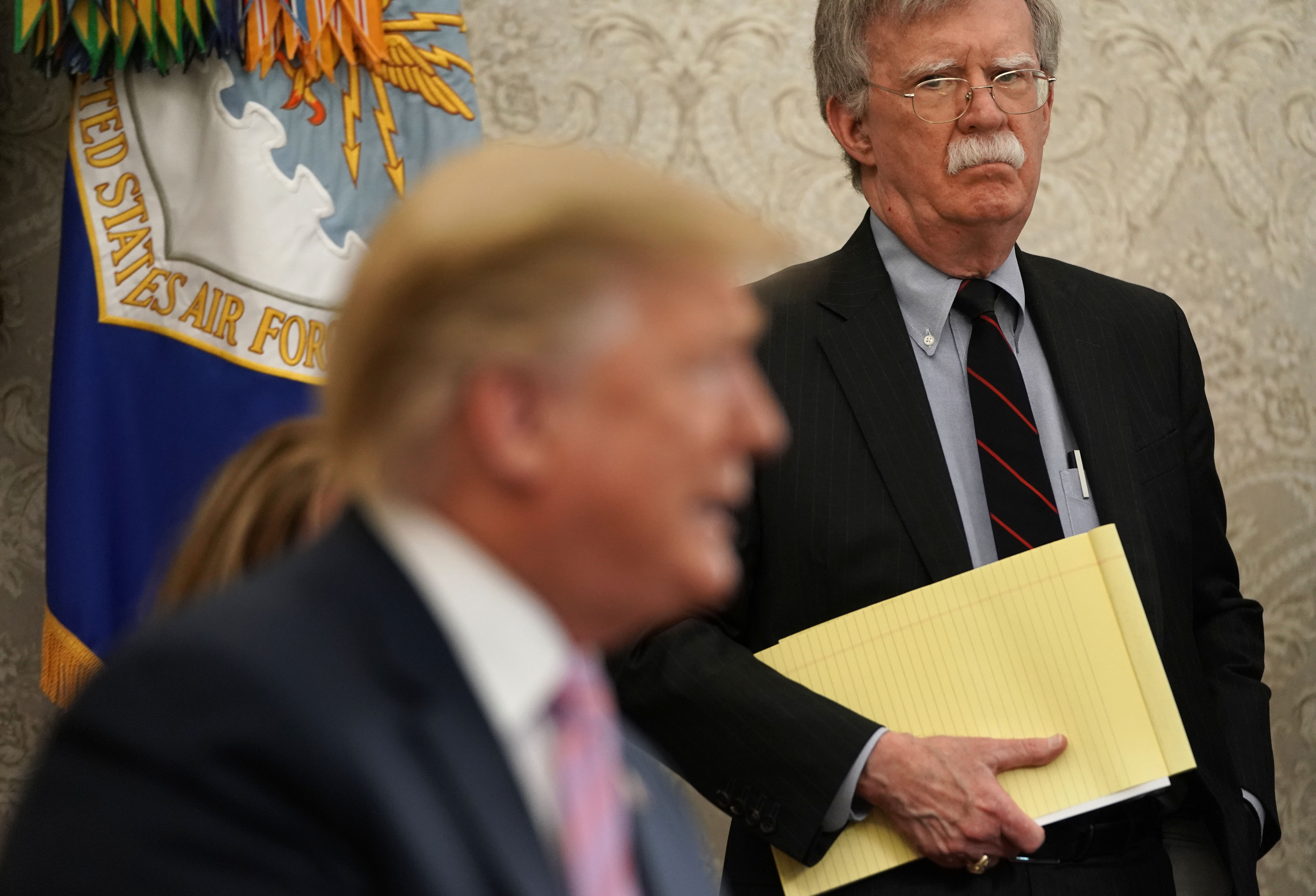 Bolton behind Trump holding a yellow notepad.