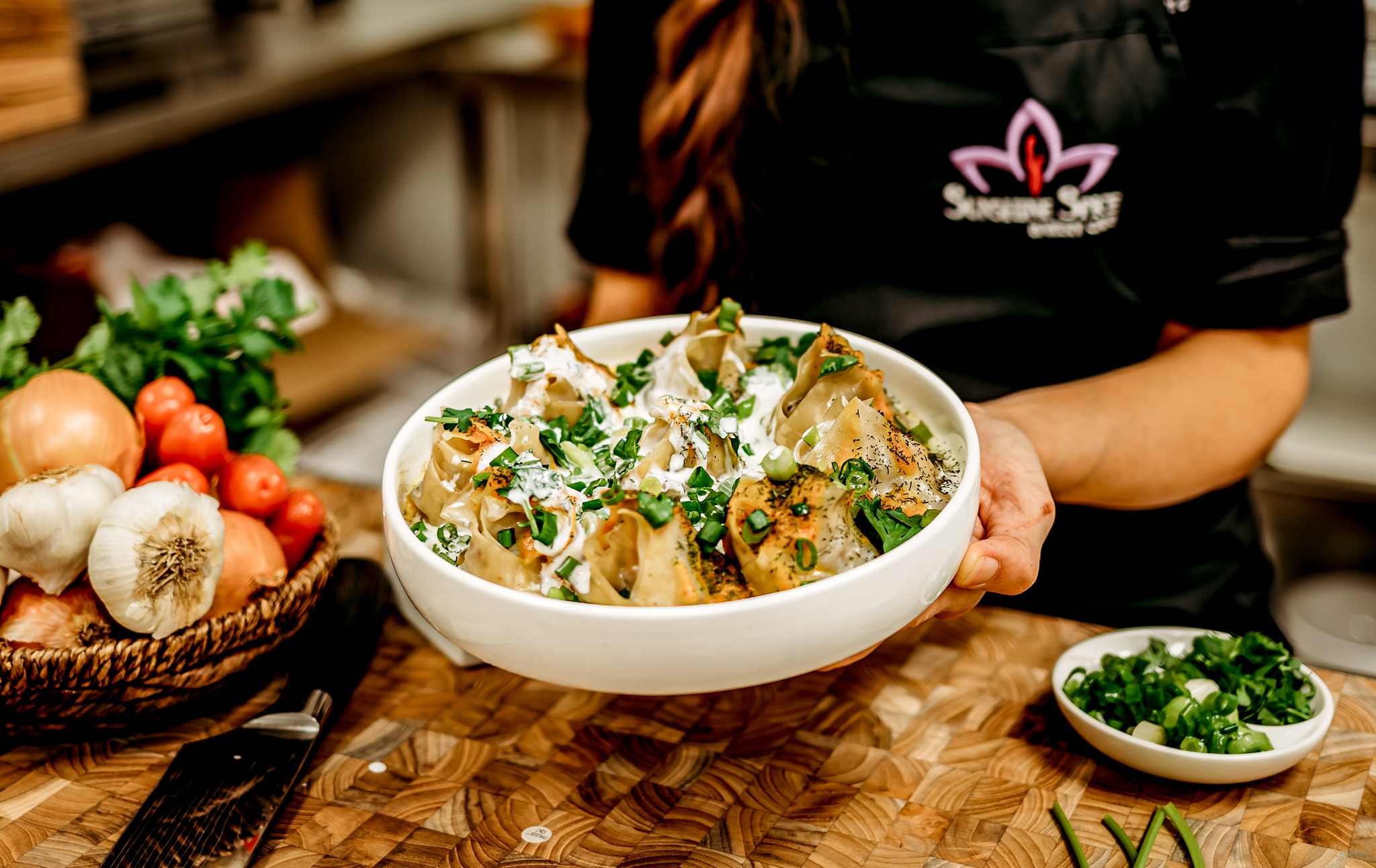 A server holds a bowl of dumplings topped with cream, shopped scallions, and herbs.
