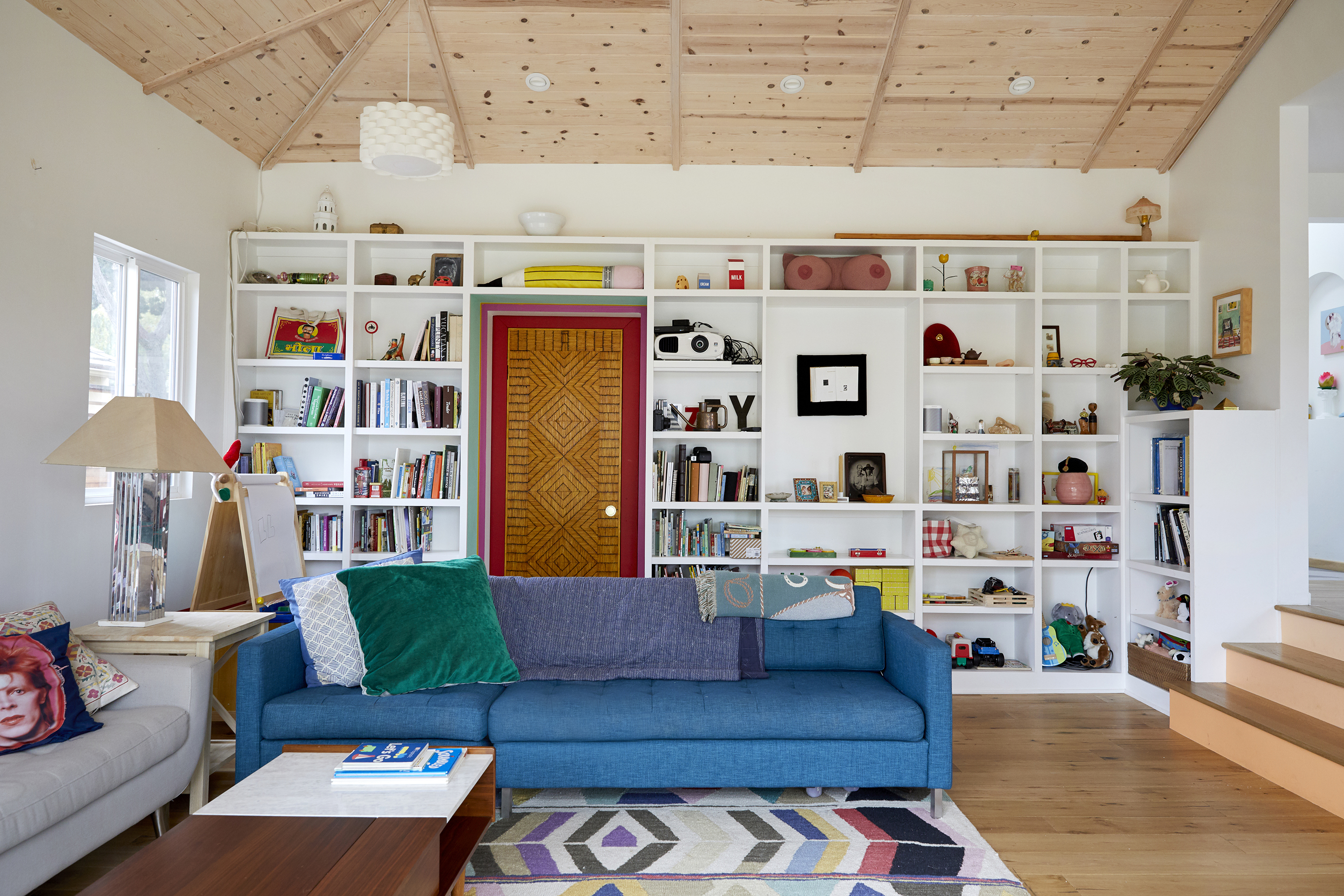 A living room with two couches. One couch is blue, the other couch is grey. On one of the walls are bookshelves full of books, and art. The bookcase surrounds a door with a painted red doorframe.
