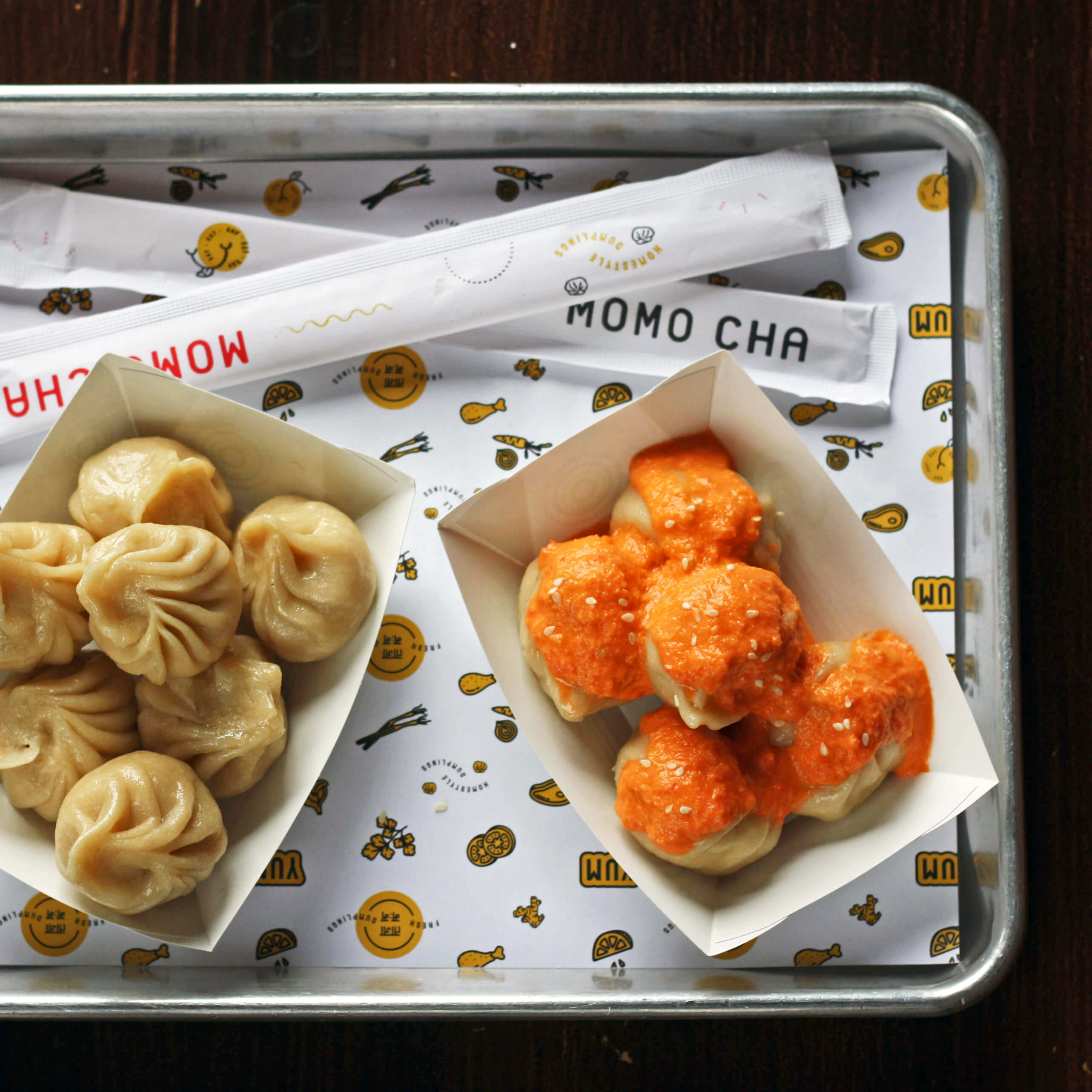 Two boats of steamed momos on a metal tray. One set of momos is topped with a bright orange chutney.