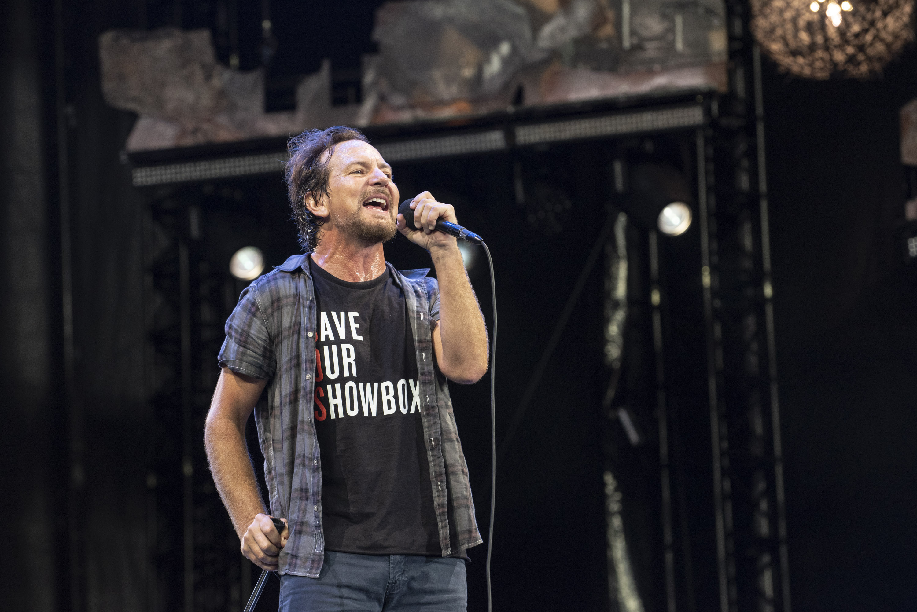 A man (Eddie Vedder) sings into a microphone while wearing a black t-shirt that says “Save Our Showbox” and a short-sleeve plaid shirt over it.