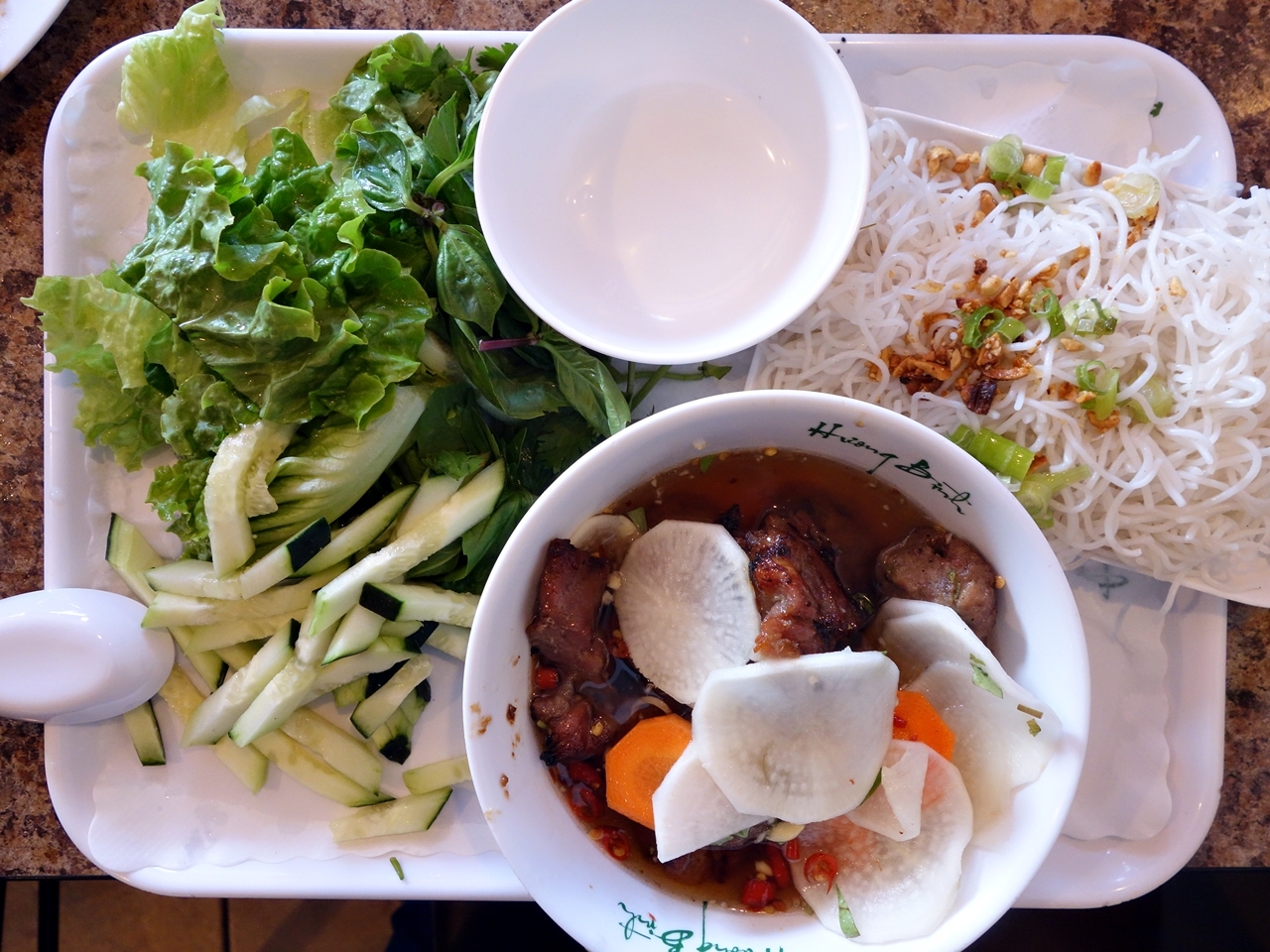 A platter with vermicelli noodles, cucumber matchsticks, shredded lettuce, grilled meat, and pickled sliced radish and carrots.