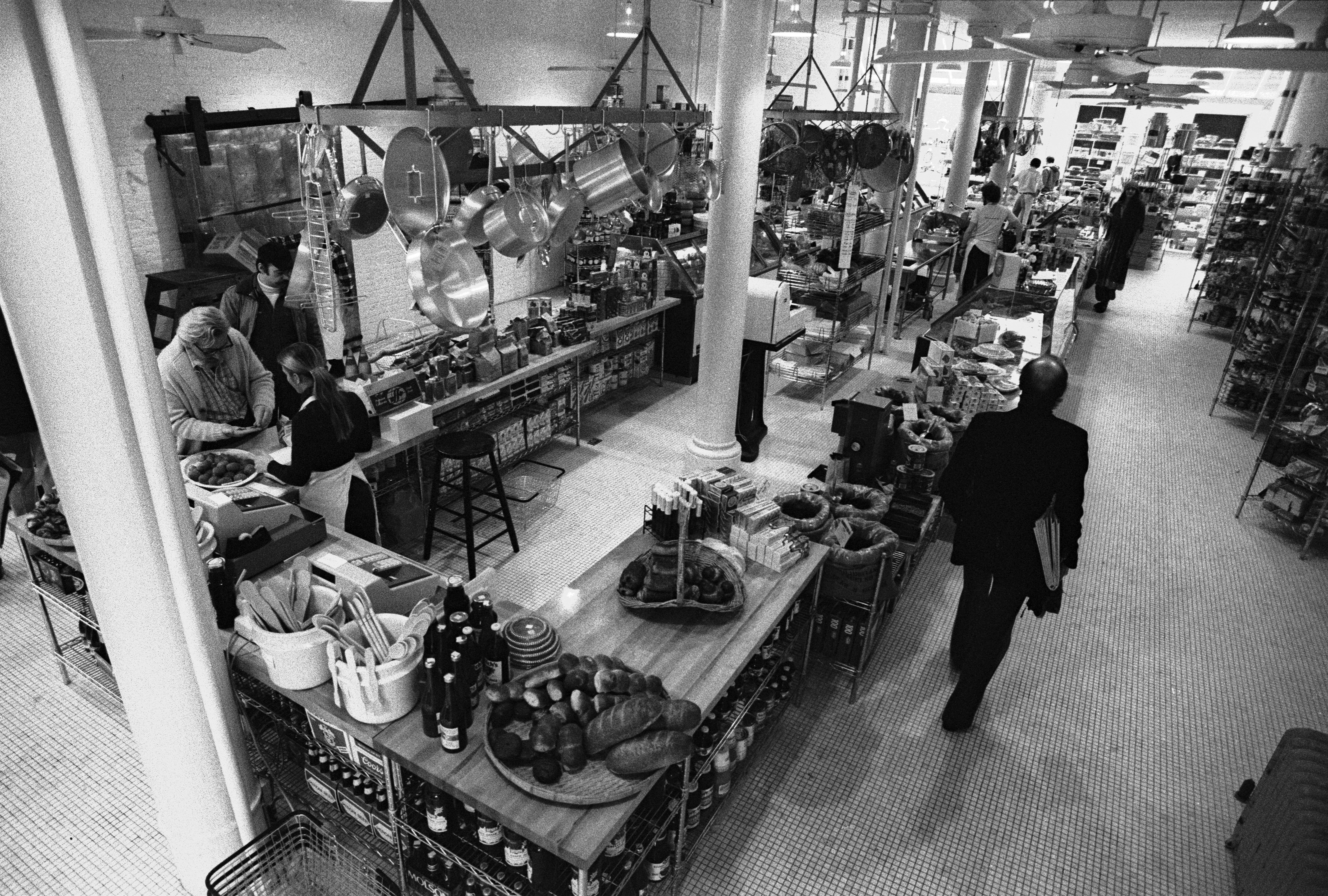 Elevated view inside the first Dean and DeLuca grocery store photographed in black and white with silver pots and pans hanging from the ceiling above a rectangular butcher block counter with baskets full of bread