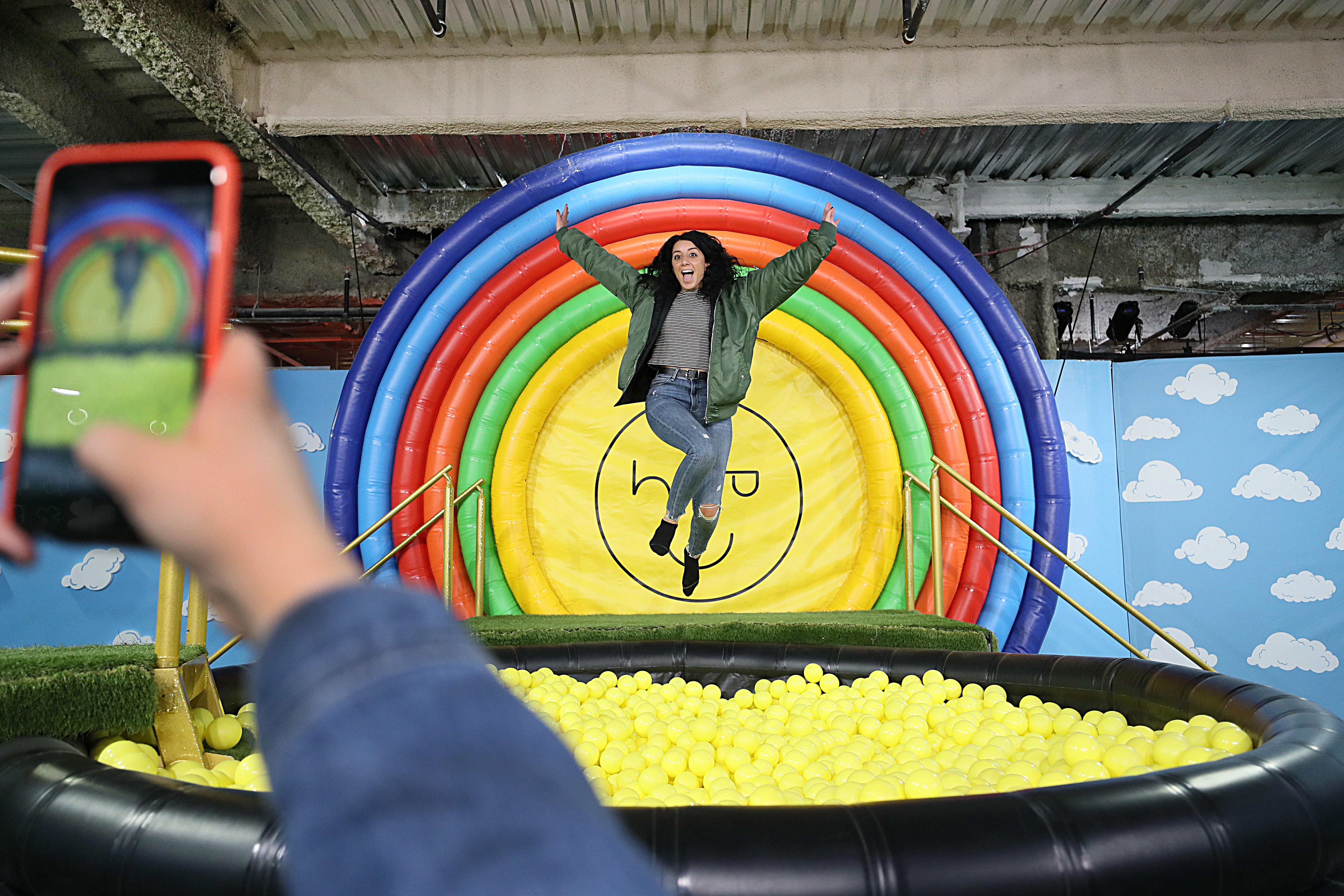 A young woman jumps into a ball pit in front of a rainbow bulls-eye design.