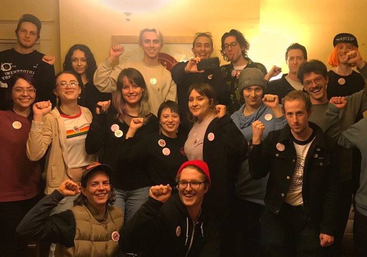 Little Big Burger employees stand together with solidarity fists. Most of them are wearing buttons for the union, standing inside a room with white walls.
