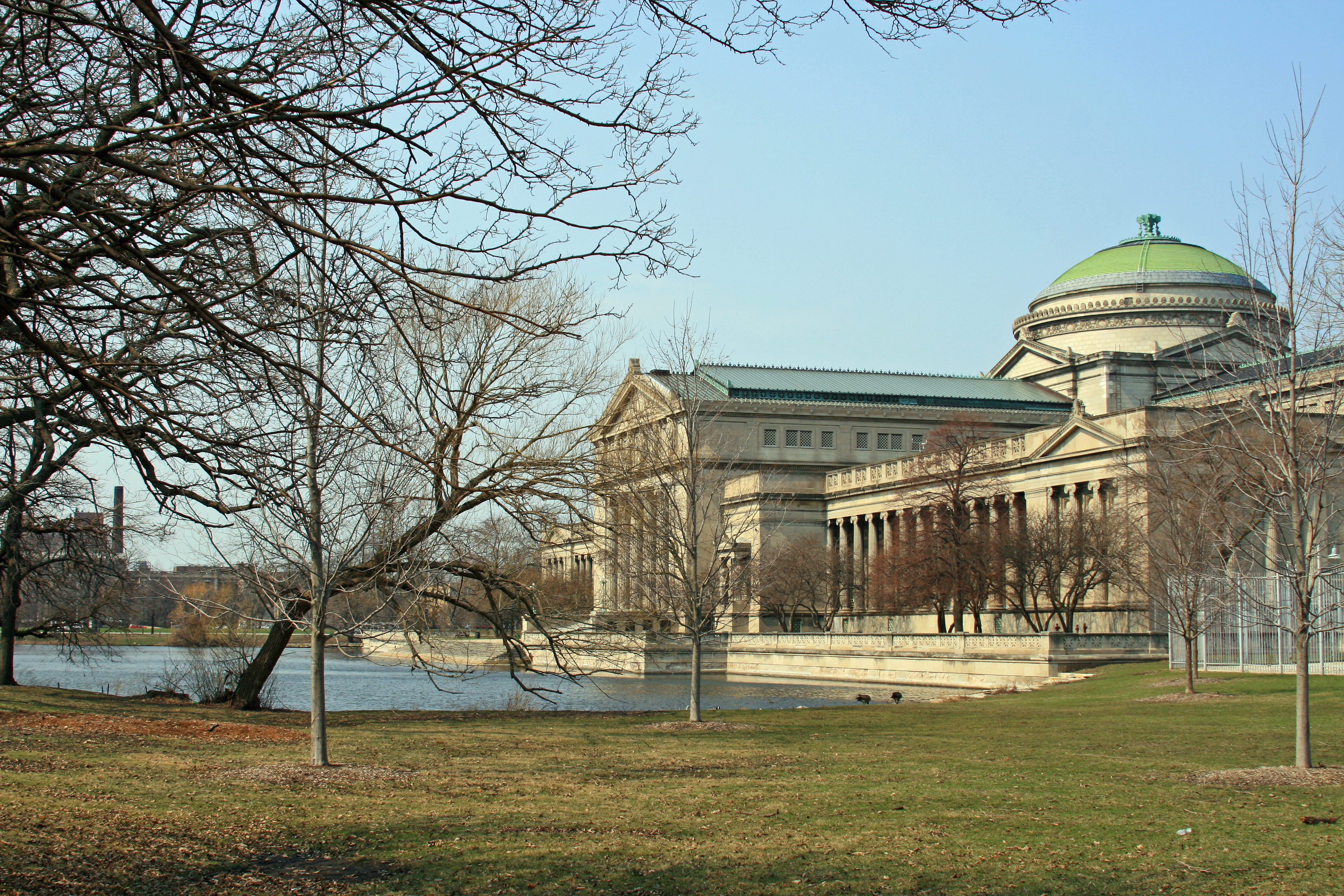 A stately building with a green dome sits in late afternoon sun overlooking a lagoon with grass and late fall trees without leaves.