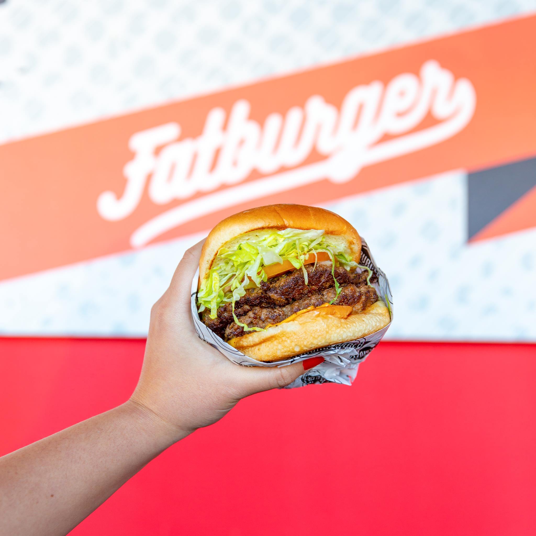 A hand holding up a semi-wrapped burger with shredded lettuce, a tomato slice, two patties, and cheese in front of a Fatburger sign
