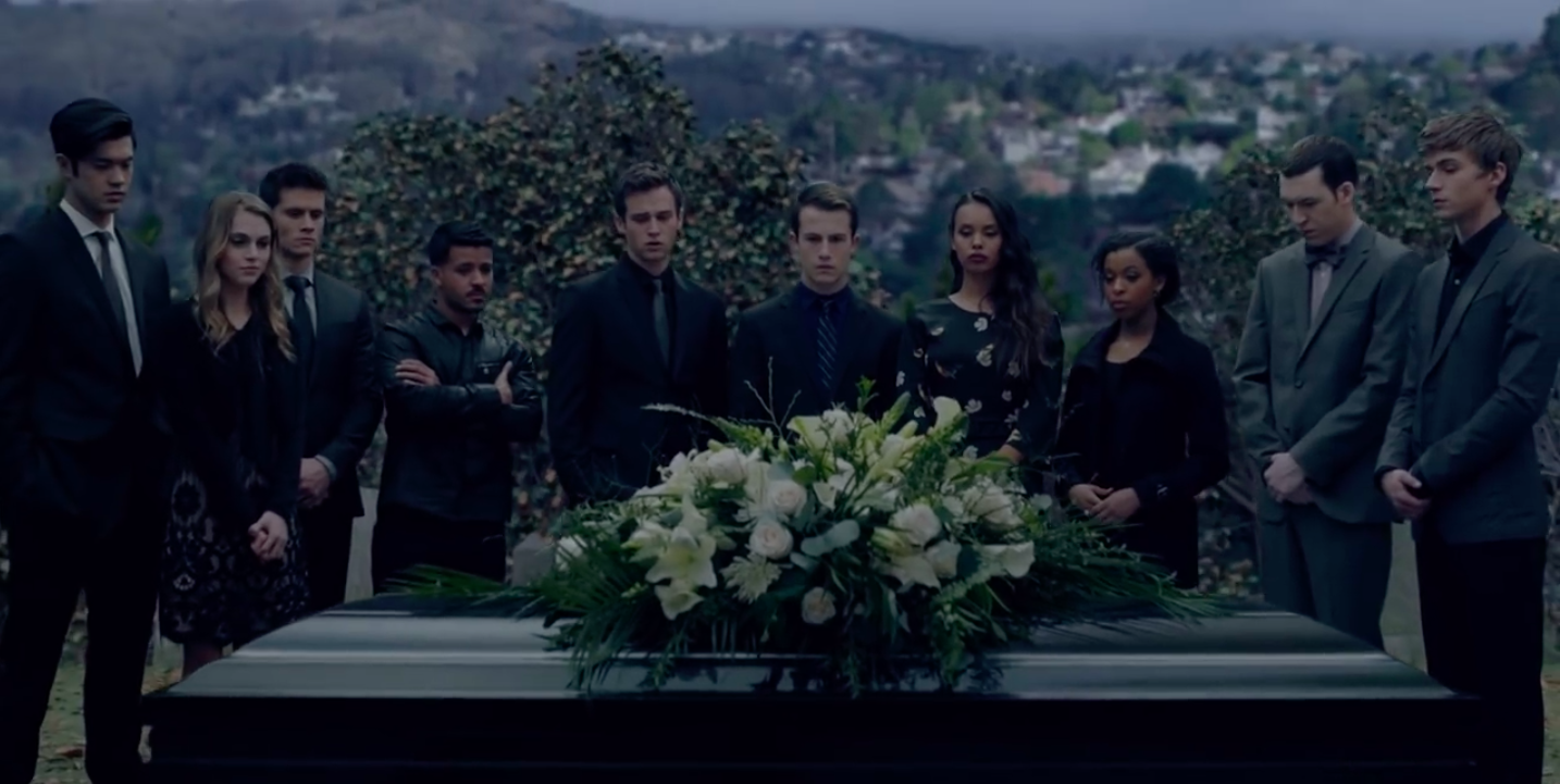 A group of students in black, formal clothes like suits and dresses stand somberly around a black coffin with white flowers resting on the lid. The sky is dark and there is a tree in the background.