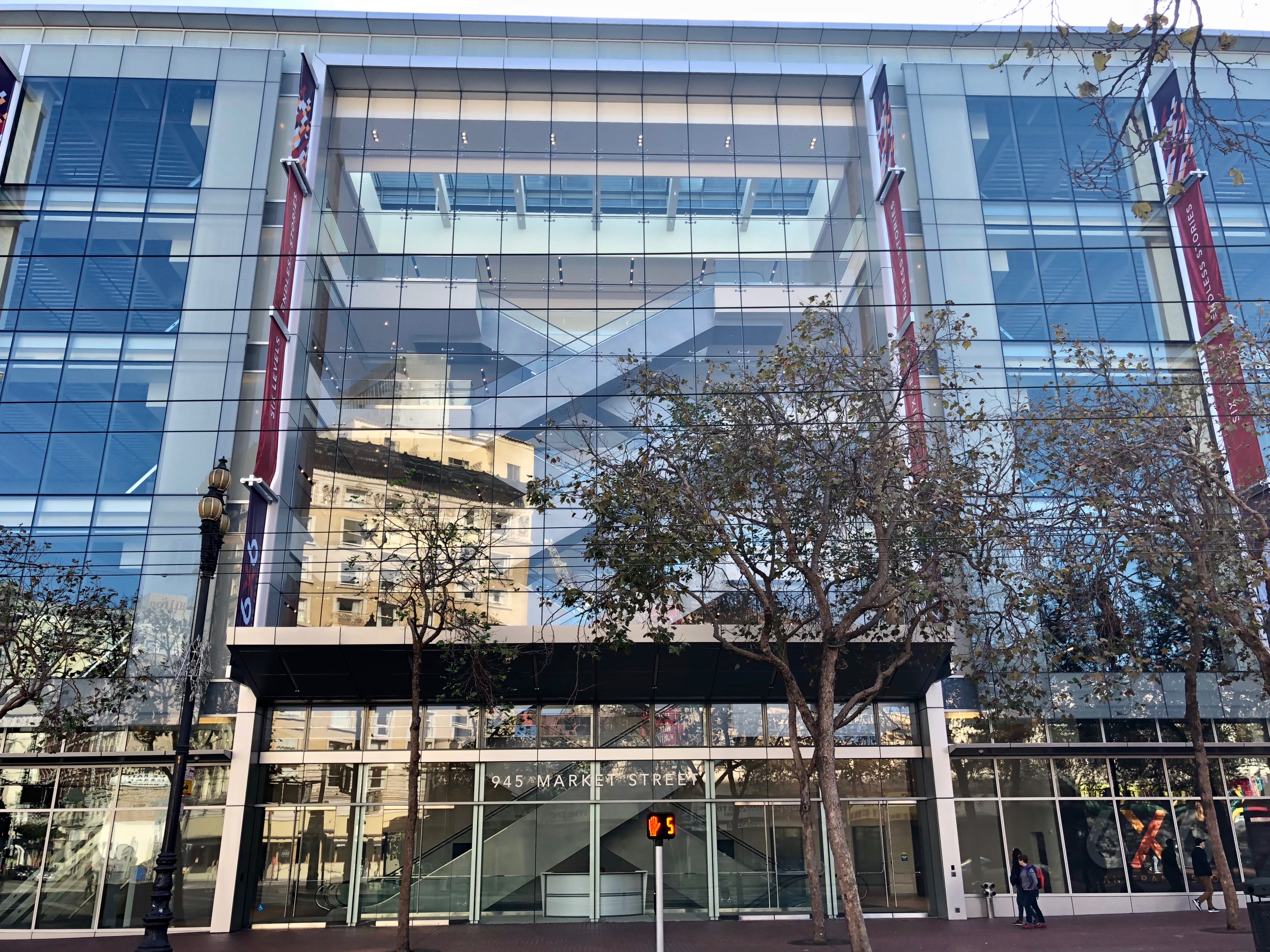 The glass facade of a giant, empty, seven-story building in San Francisco, with criss-crossing escalators visible from the street.